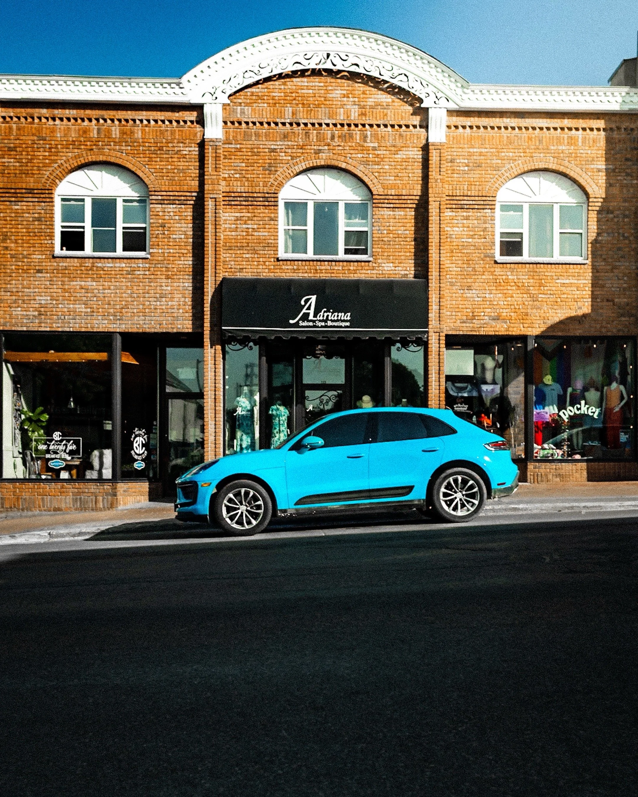 A brick building with large windows and a black awning that reads 'Adriana Salon Spa Boutique.' A blue car is parked in front of the building on a dark asphalt street, and a clear blue sky is above.