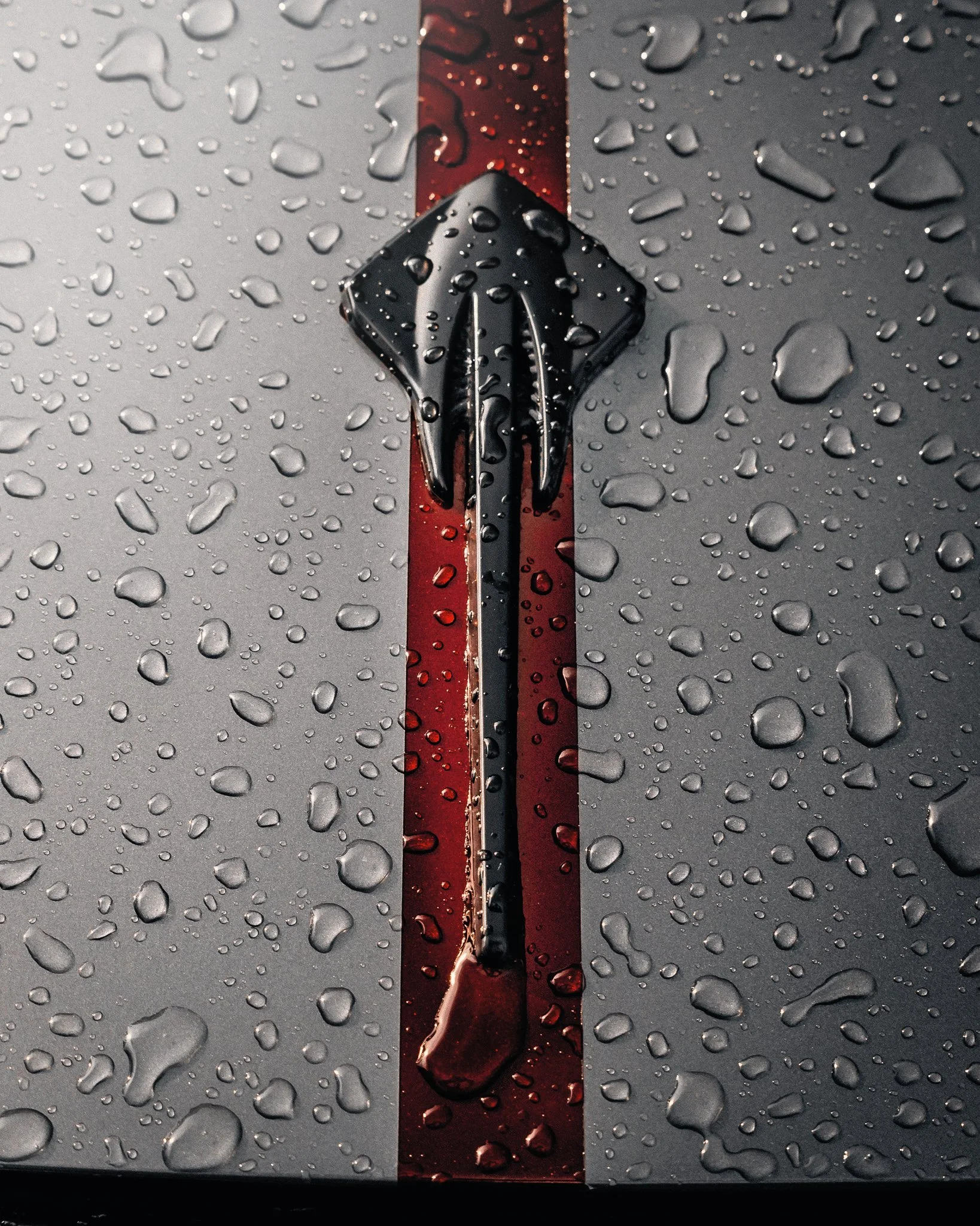 A close-up of a metallic surface with rain droplets, featuring a black and red fountain pen resting on a red stripe that runs vertically down the middle of the surface.
