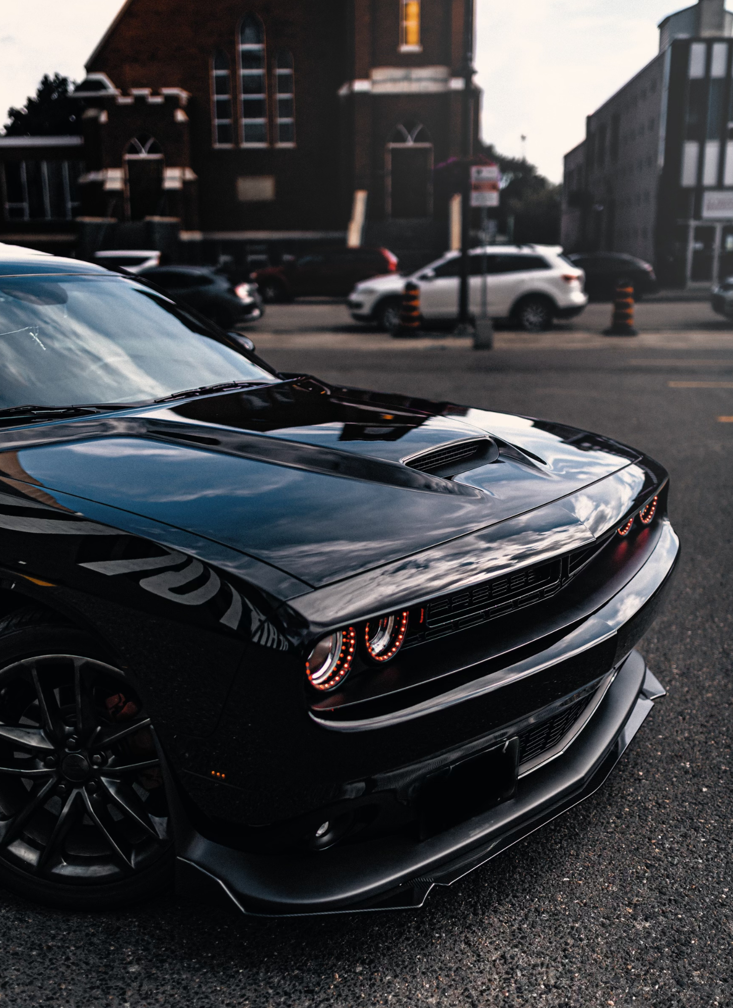 Close-up of the front of a black sports car with illuminated headlights, parked on a street, with other vehicles and brick buildings in the background.