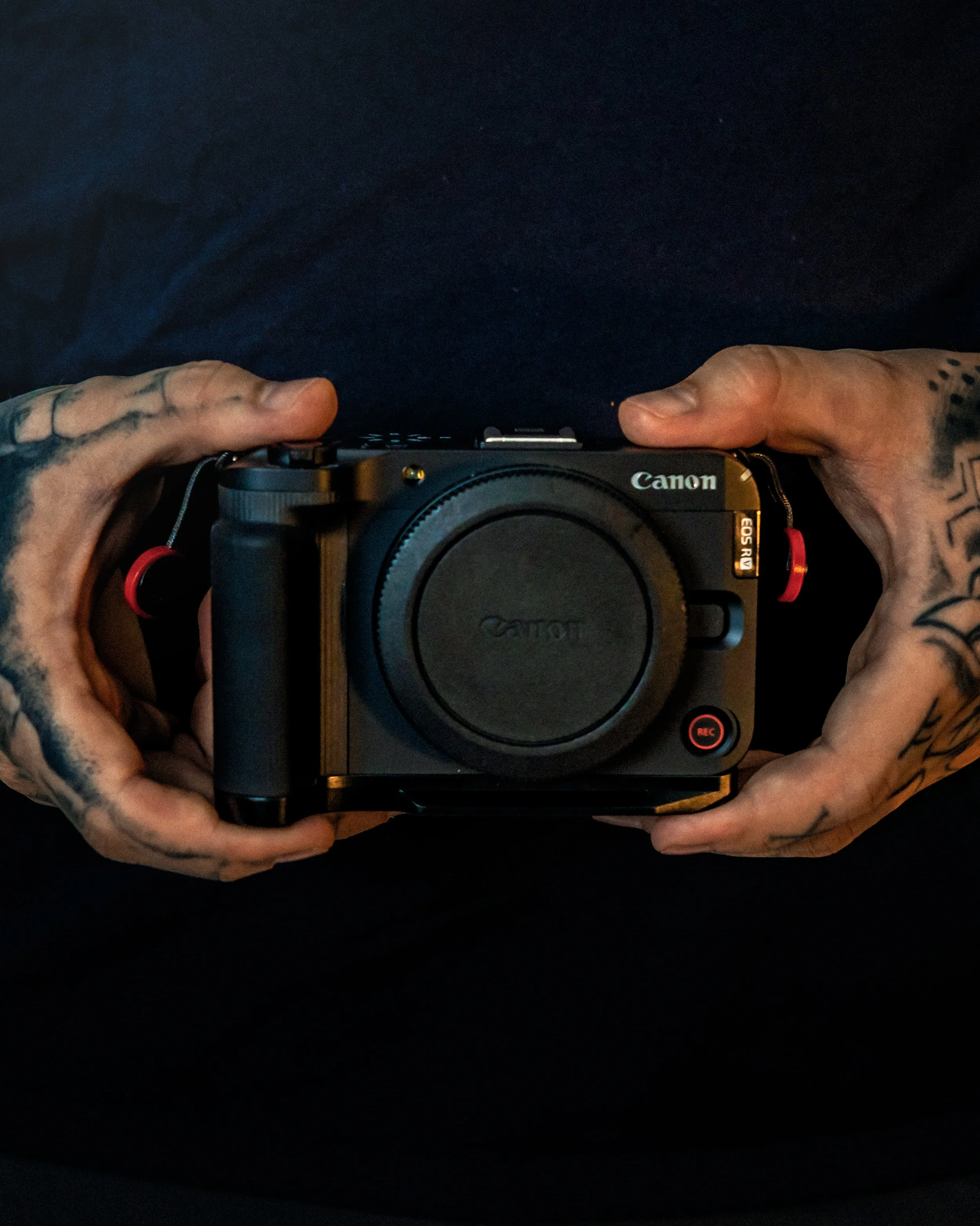 A person with tattooed hands holding a Canon camera with both hands, positioned above a black surface, with the camera facing downward.
