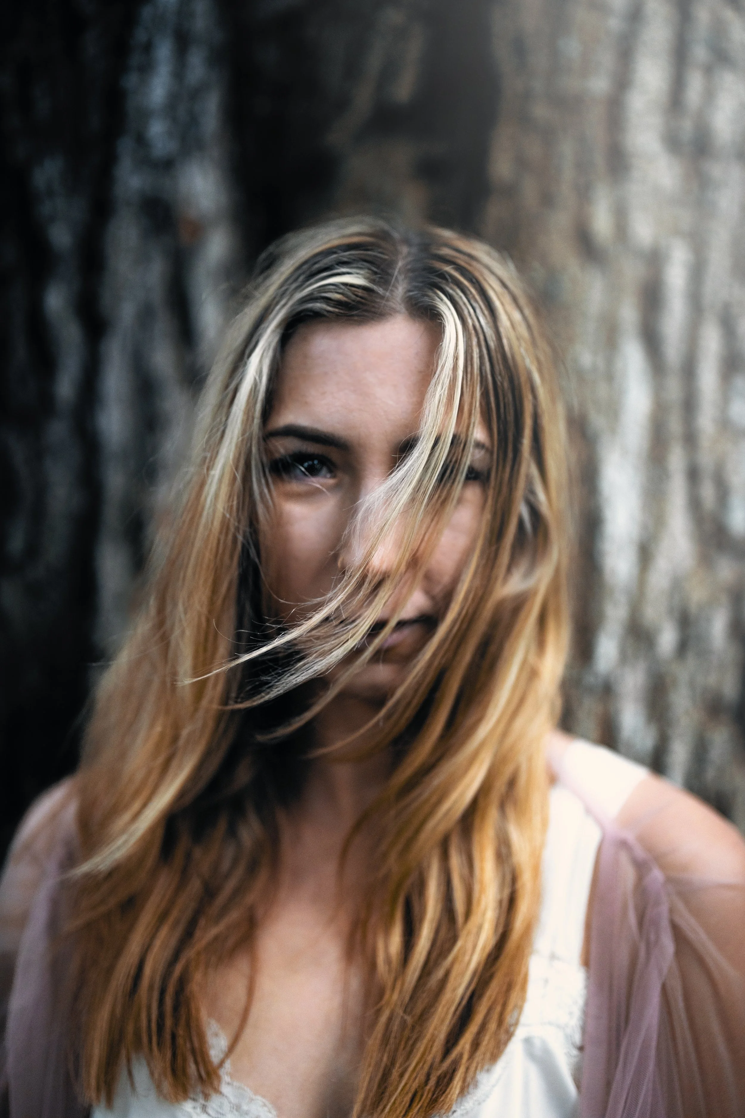 A woman with long, wavy hair with blonde highlights, smiling with teeth showing, wearing a sleeveless white top, standing outdoors near a large tree trunk.