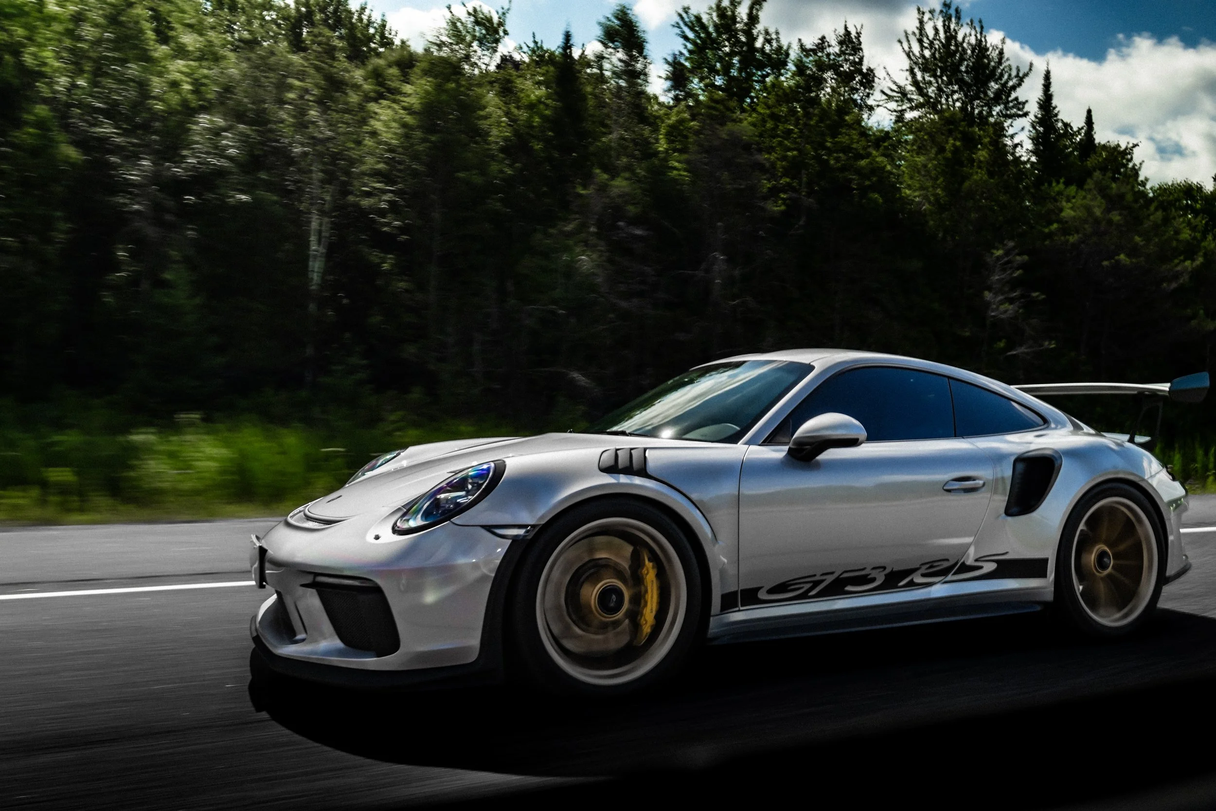 A silver sports car with black and yellow wheels driving on a highway with a forest background and partly cloudy skies.