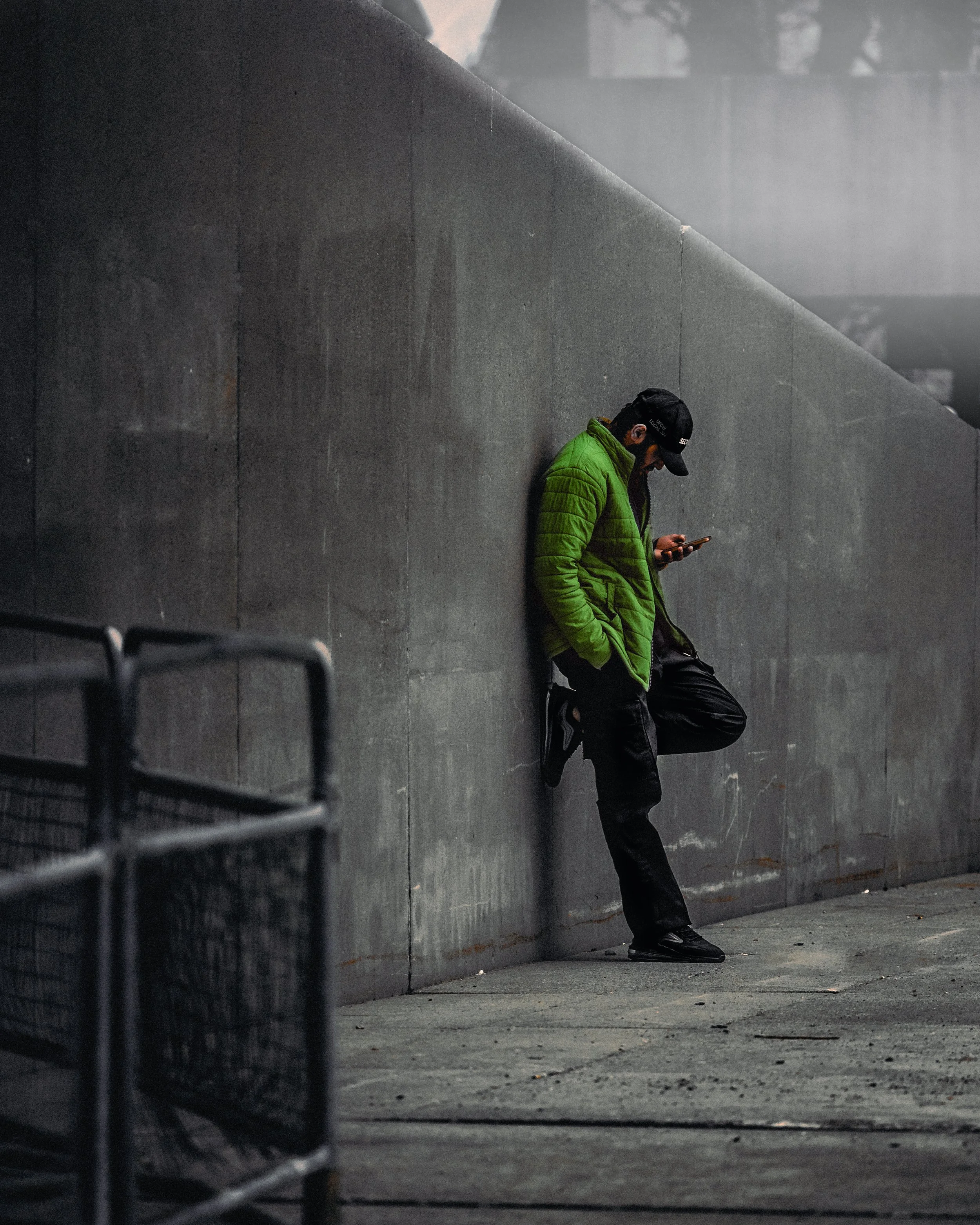 A man in a bright green jacket and black cap leaning against a dark gray concrete wall, looking at his phone. There is an unoccupied bench partially visible in the foreground.