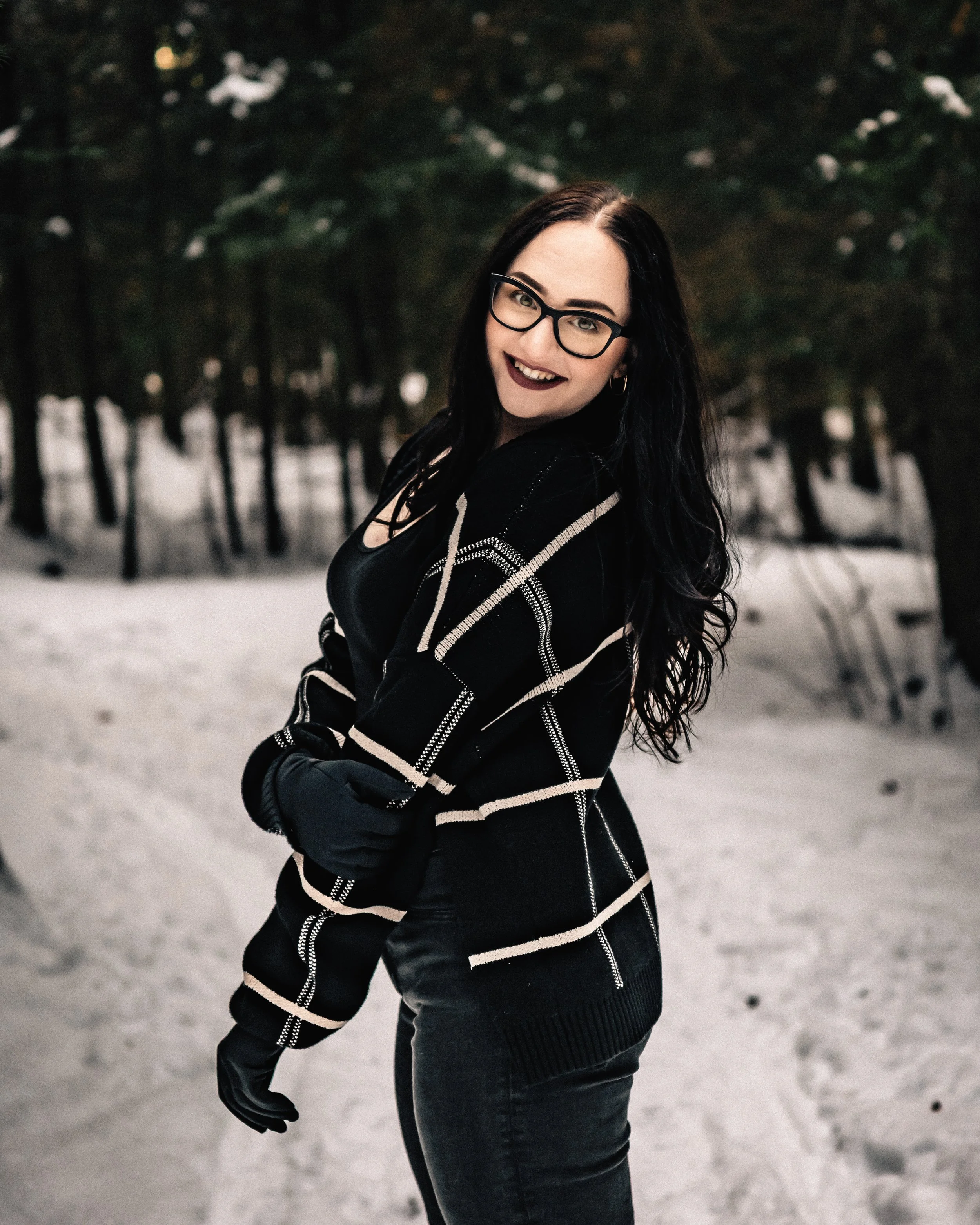 A woman with long black hair, glasses, and dark lipstick smiling outdoors in a snowy, wooded area, wearing a black and white striped jacket and black gloves.
