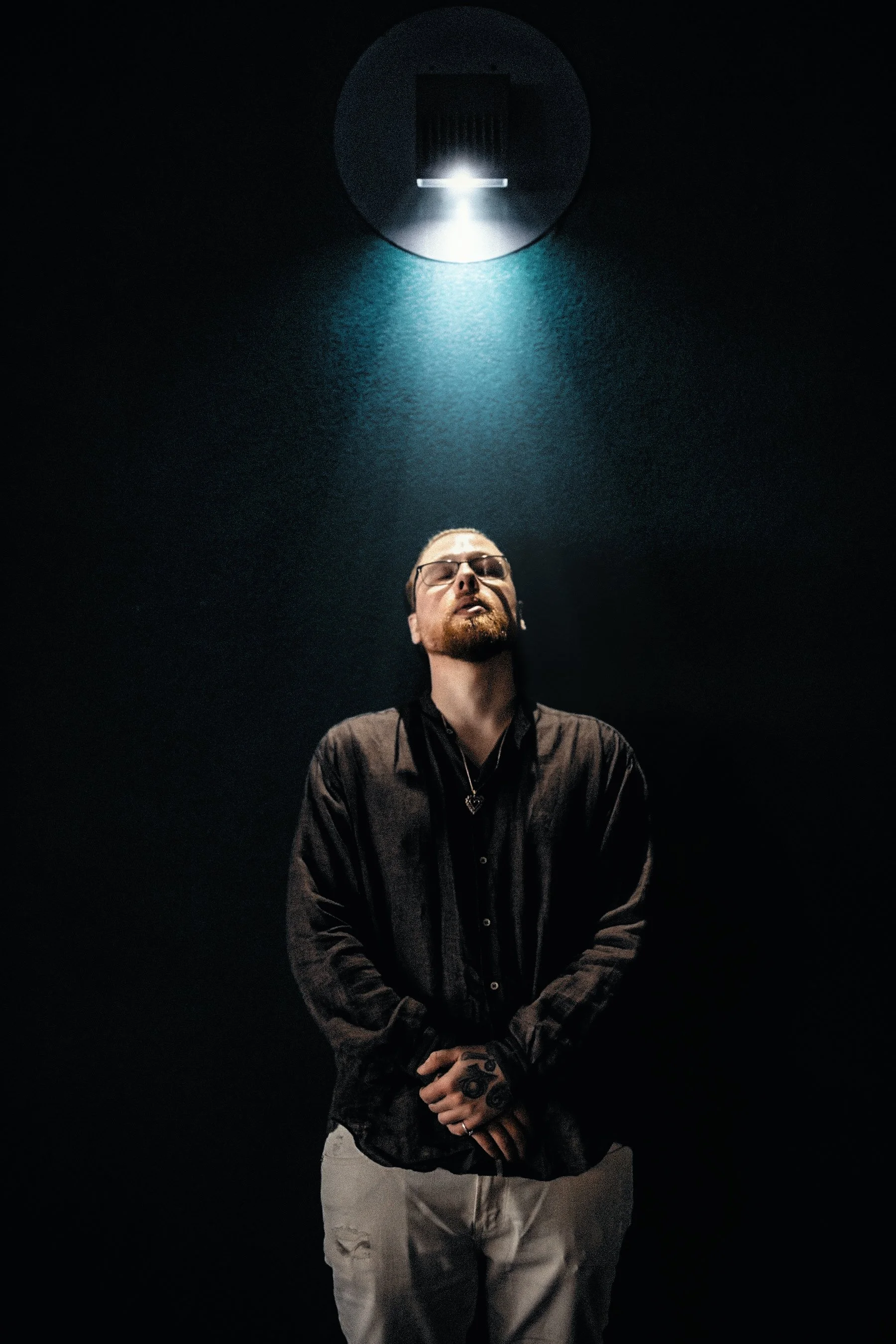 A man with glasses and a beard standing in front of a dark background, beneath a large, circular light fixture shining downward.