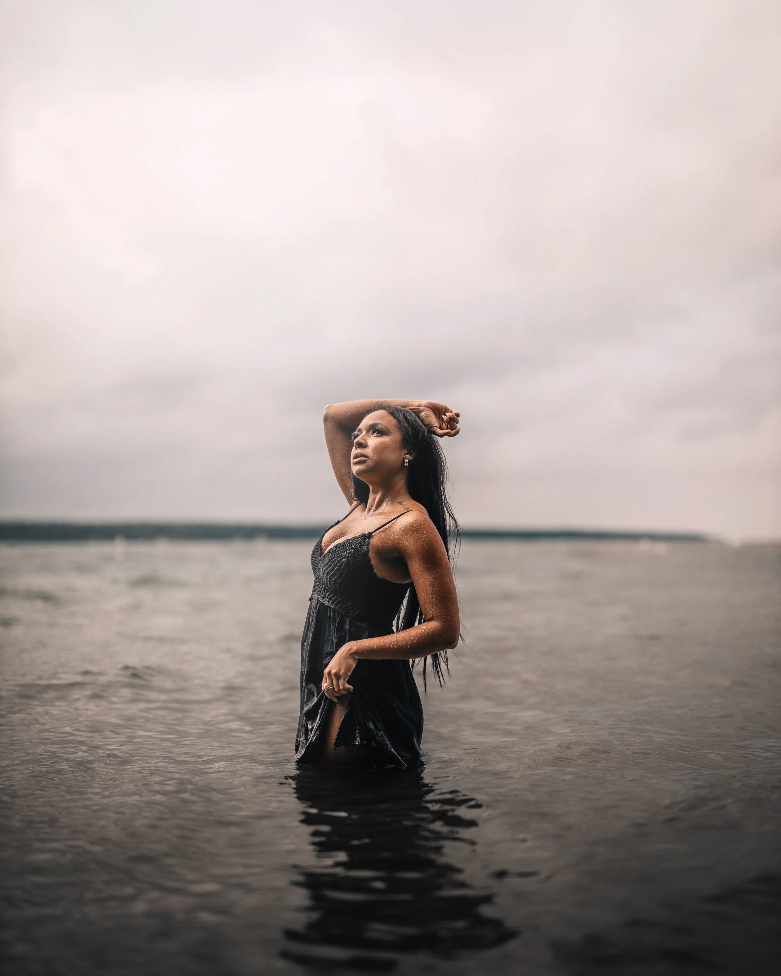 Woman standing in the water at the beach with her hand behind her head, looking thoughtful against a cloudy sky.