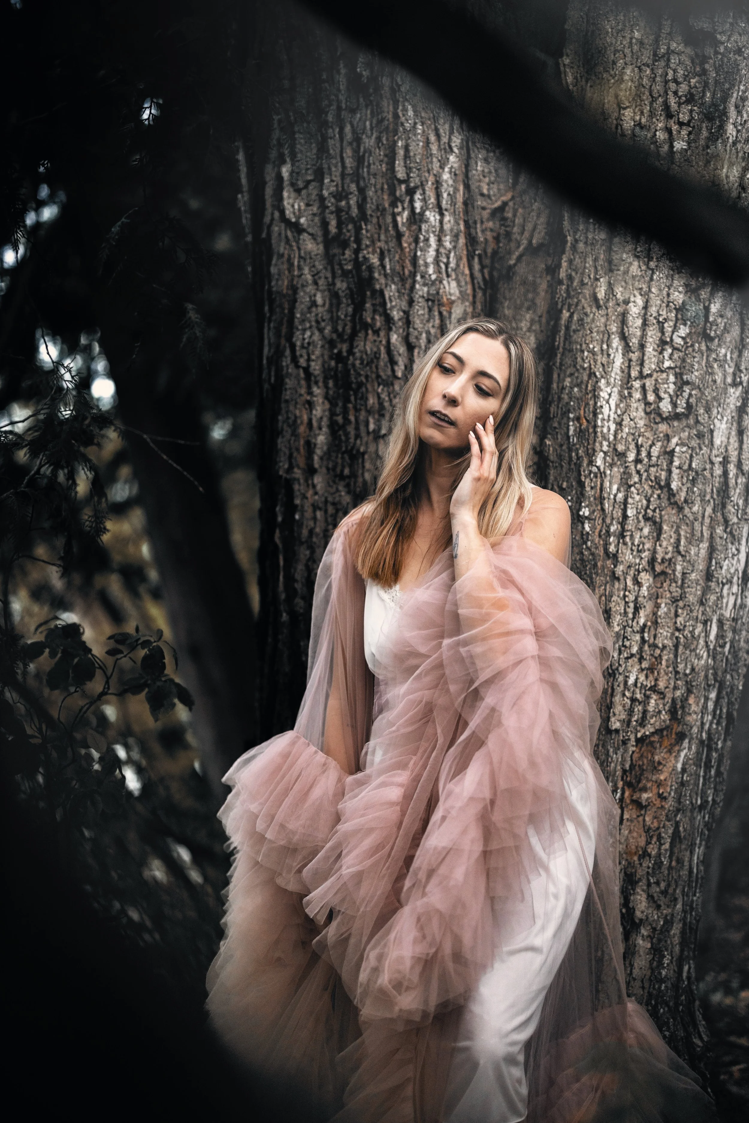 A woman with long blonde hair, wearing a flowing, sheer pink dress, stands against a large tree in a forest, touching her face with one hand, looking thoughtfully to the side.