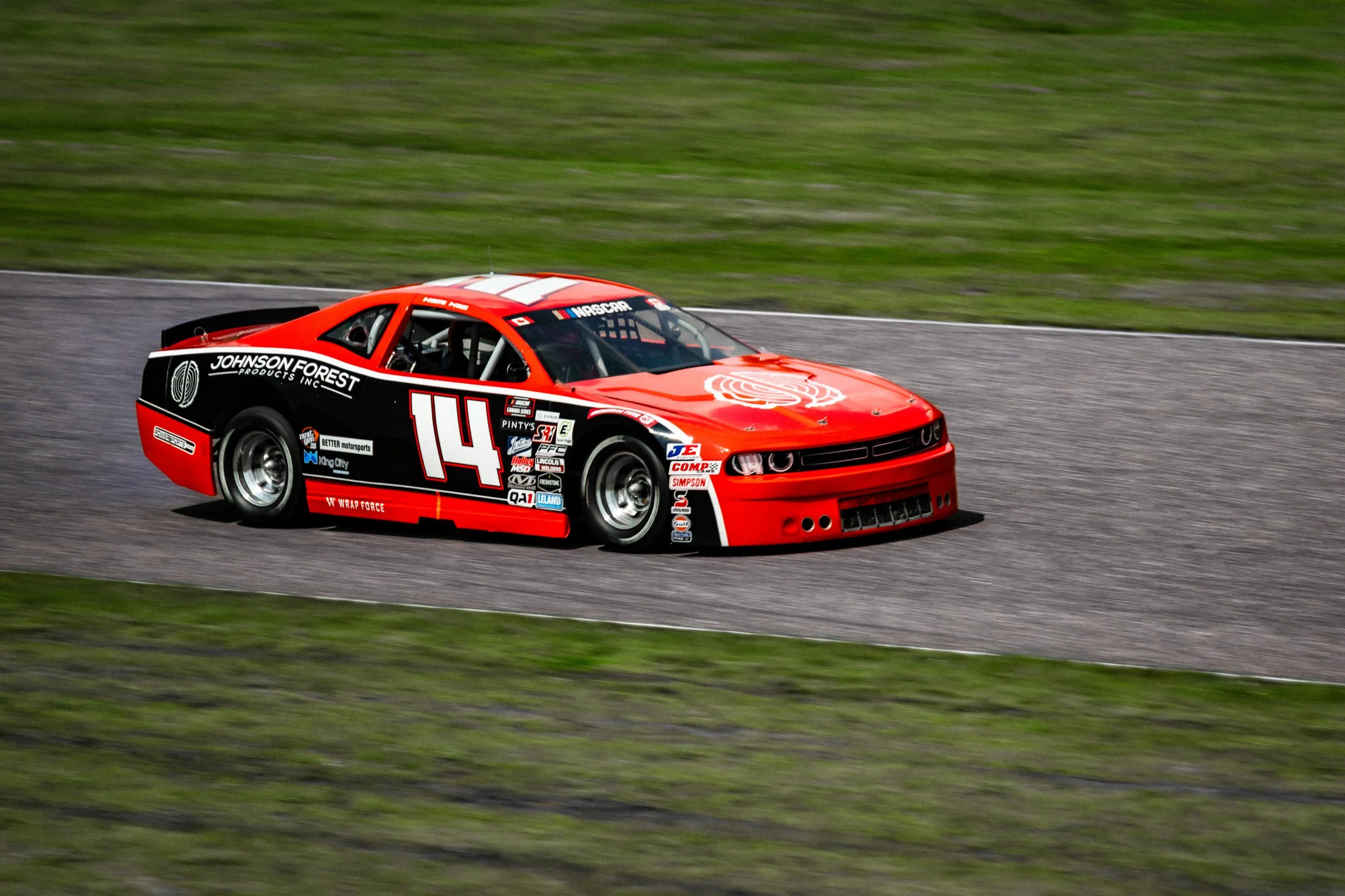 A red race car with number 14 and sponsorship logos driving on a racetrack with grass in the background.