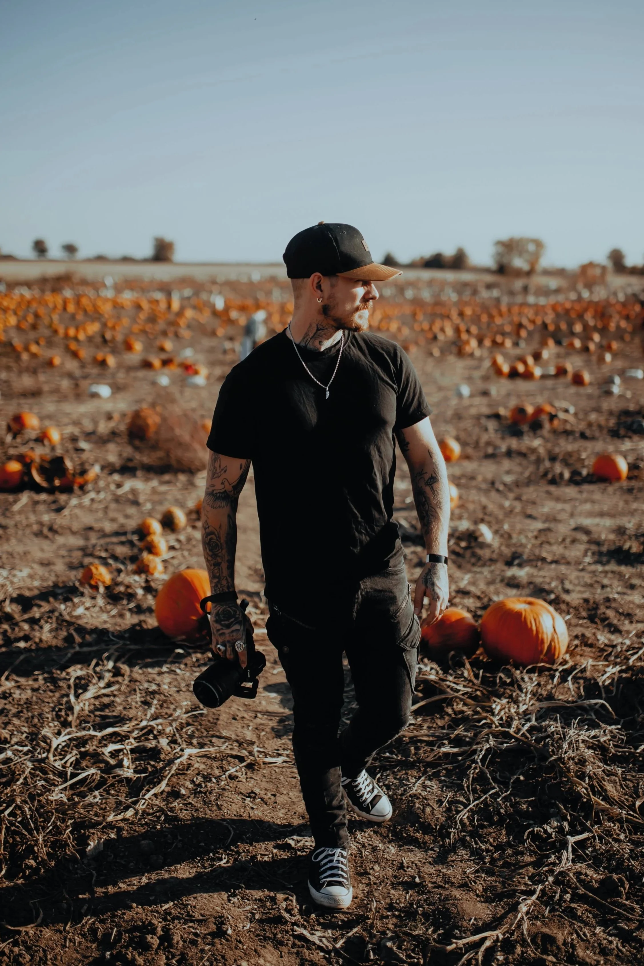 A man with tattoos wearing a black t-shirt and black cap walking through a pumpkin patch during daytime, holding a camera.