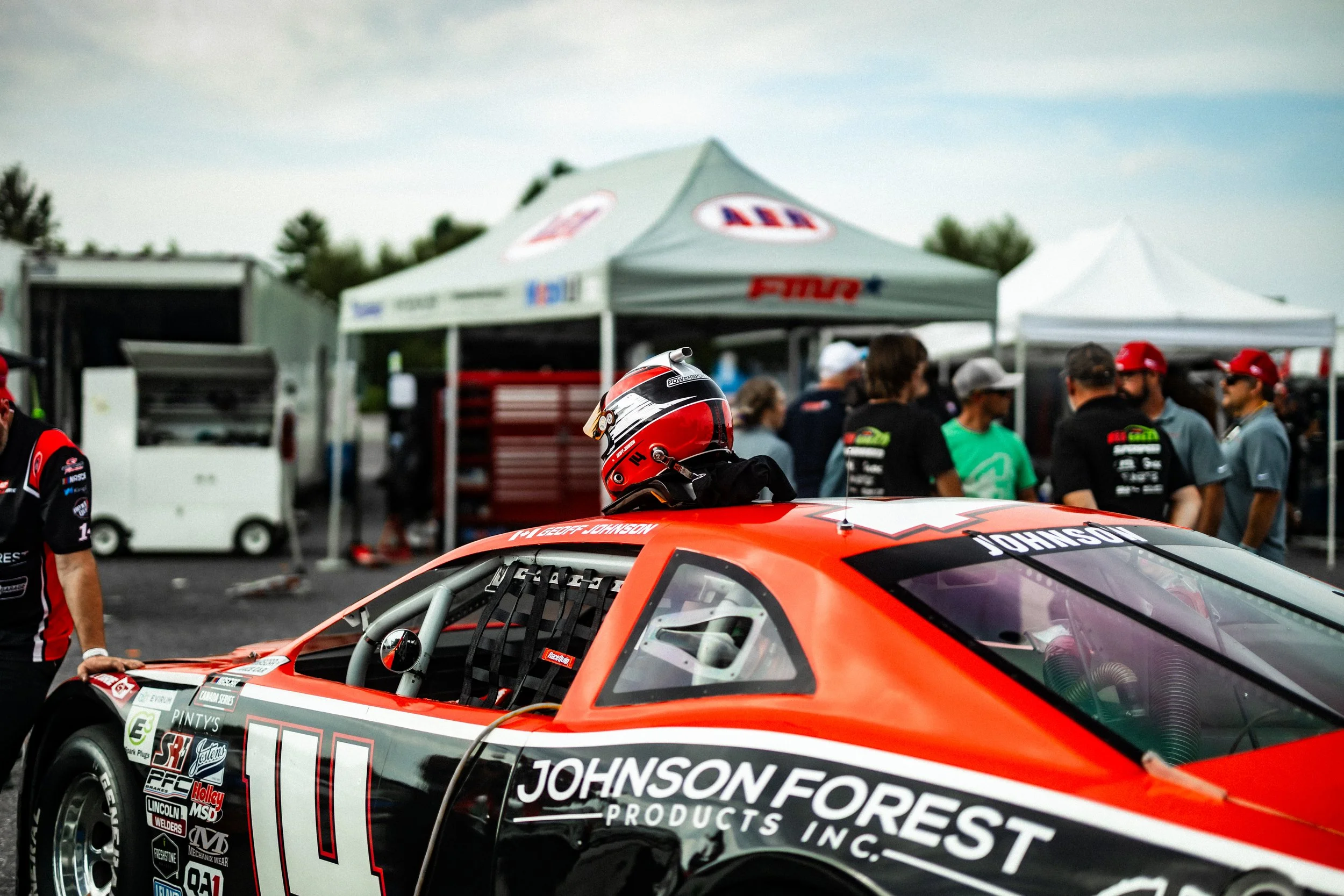 Race car with numerous sponsor decals, red and black livery, and the name 'Johnson Forest Products Inc.' on the side, in a racing paddock area with team members and tents in the background.
