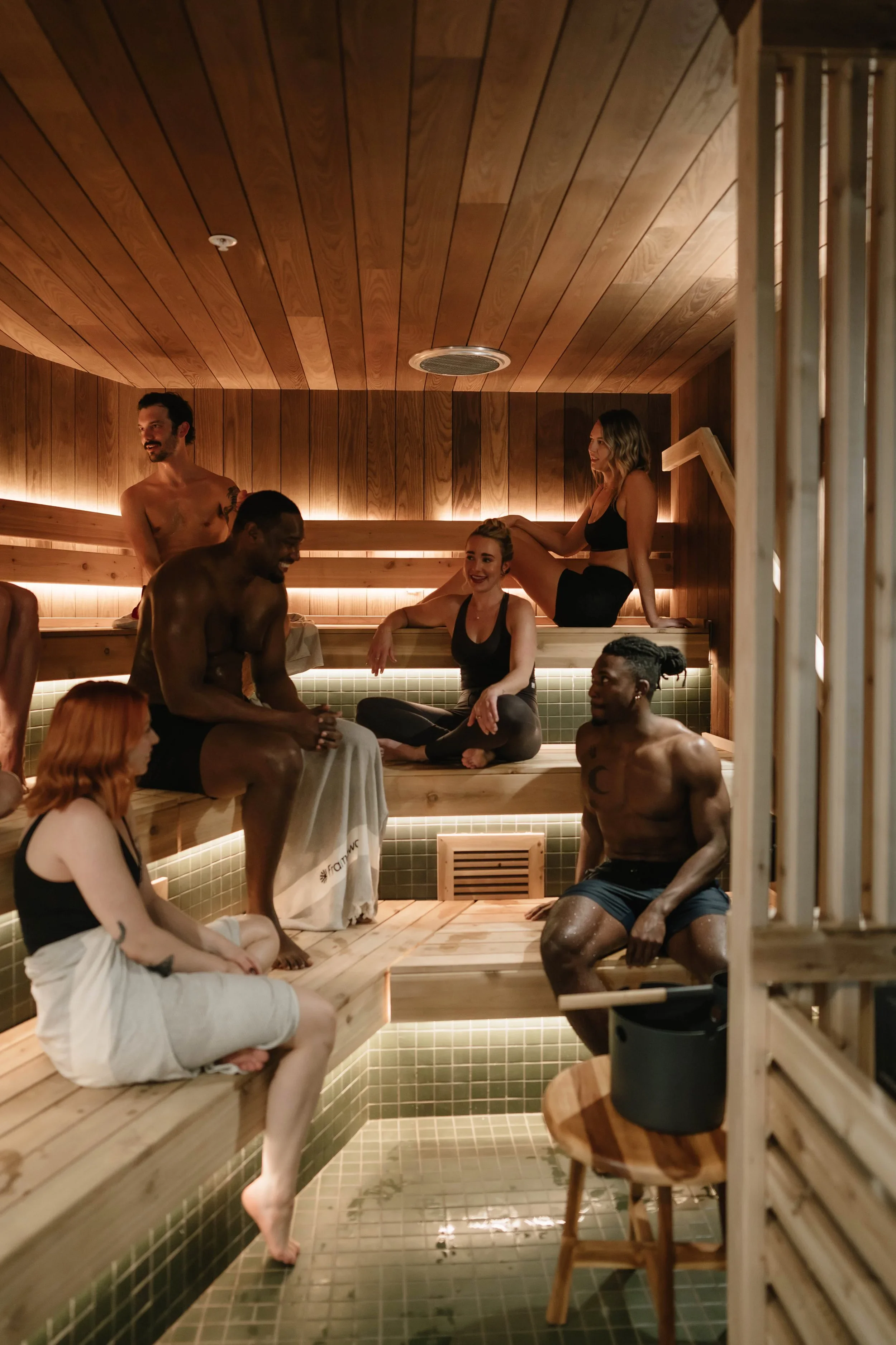 People relaxing and socializing in a wooden sauna or steam room with tiled floor and warm lighting.