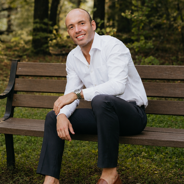 A man smiling and sitting on a park bench in a wooded area, wearing a white shirt, dark pants, and brown shoes.