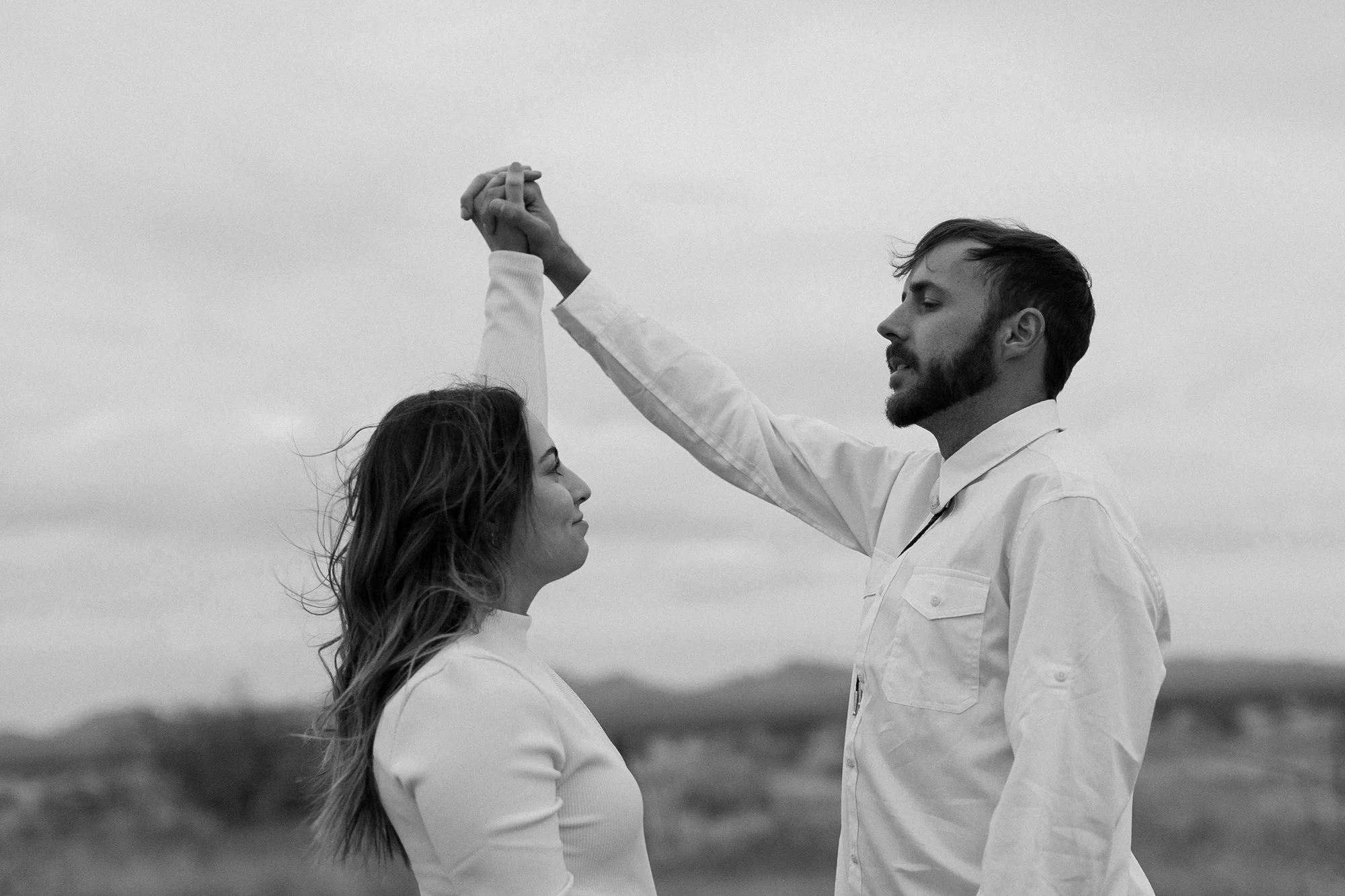 A black-and-white photo of a man and a woman holding hands with their arms raised, standing outdoors with a blurred natural background.