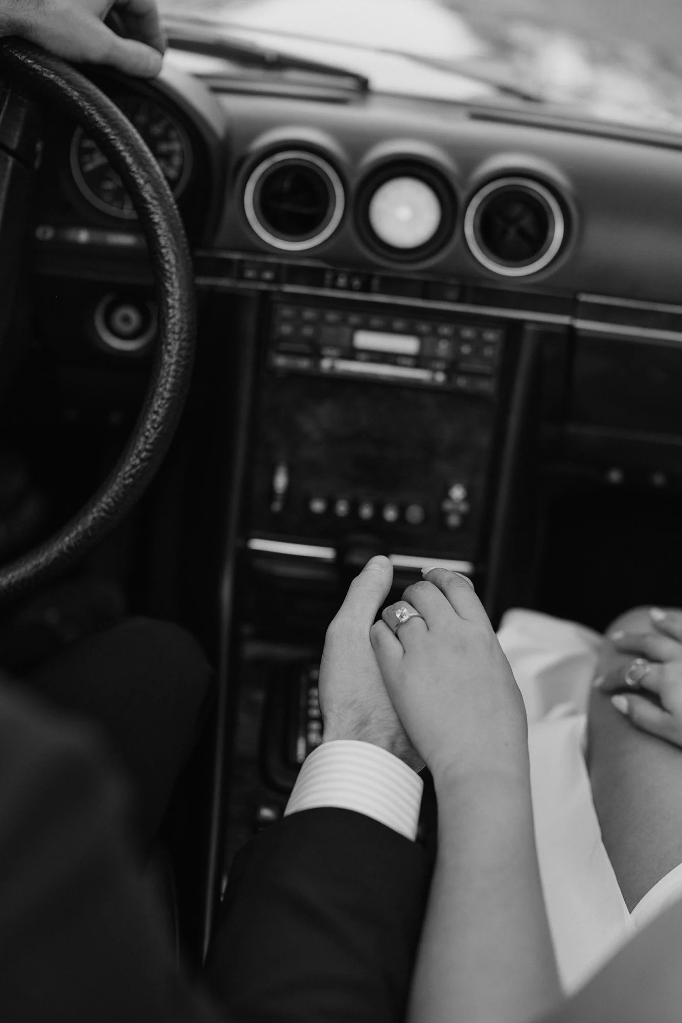 Black and white photo of a person's hand holding a child's hand inside a car. The focus is on their hands, with the car's dashboard and steering wheel visible in the background.