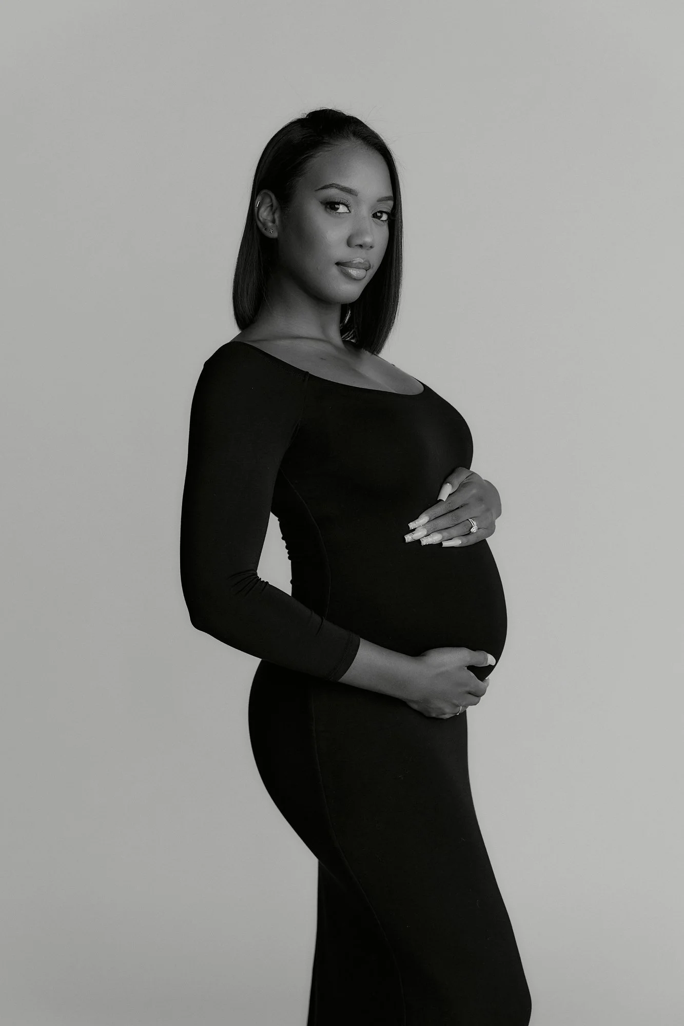 Black and white photo of a pregnant woman in a form-fitting black dress, standing sideways with her hands on her belly, looking at the camera against a plain background.
