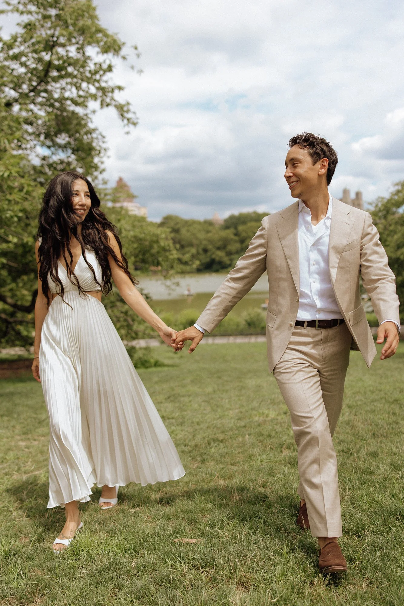 A happy couple holding hands and walking outdoors in a park, smiling at each other, with trees and a river in the background.