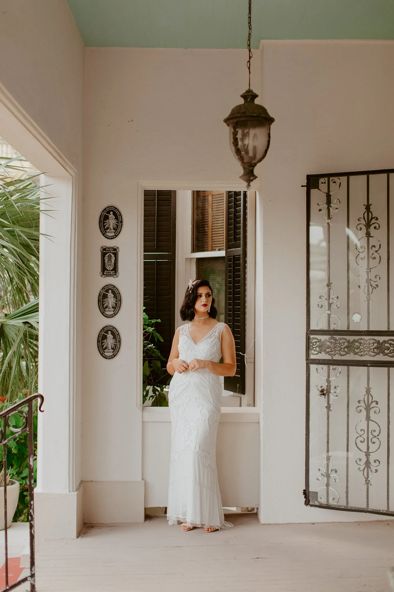 A woman in a white wedding dress standing on a porch near a window with open shutters, surrounded by plants and decorative wall plaques.