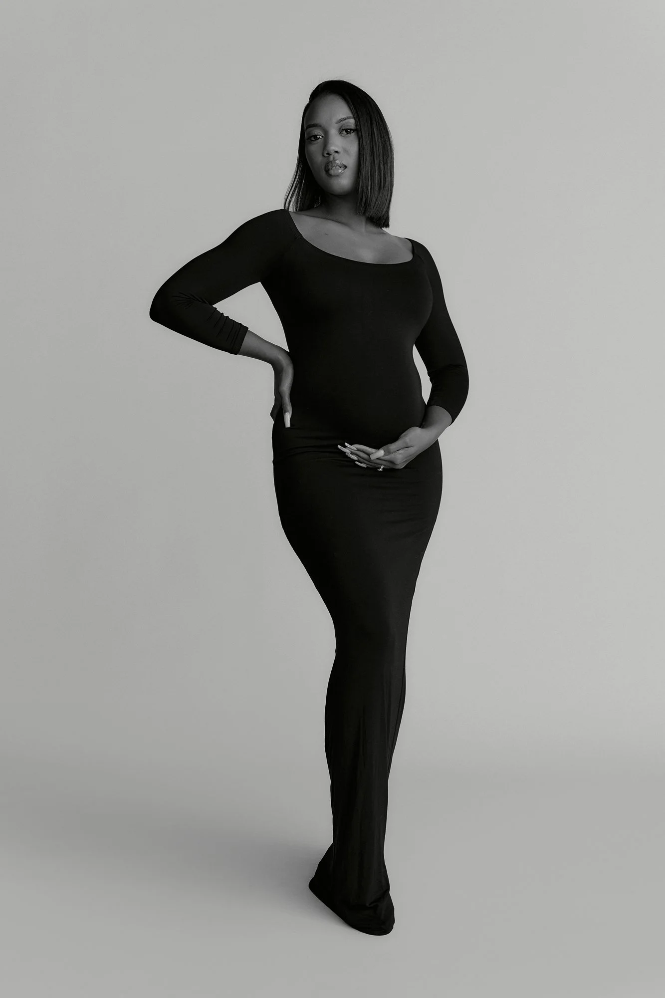 A woman in a fitted black dress with long sleeves standing with one hand on her hip and the other resting on her stomach against a plain background.