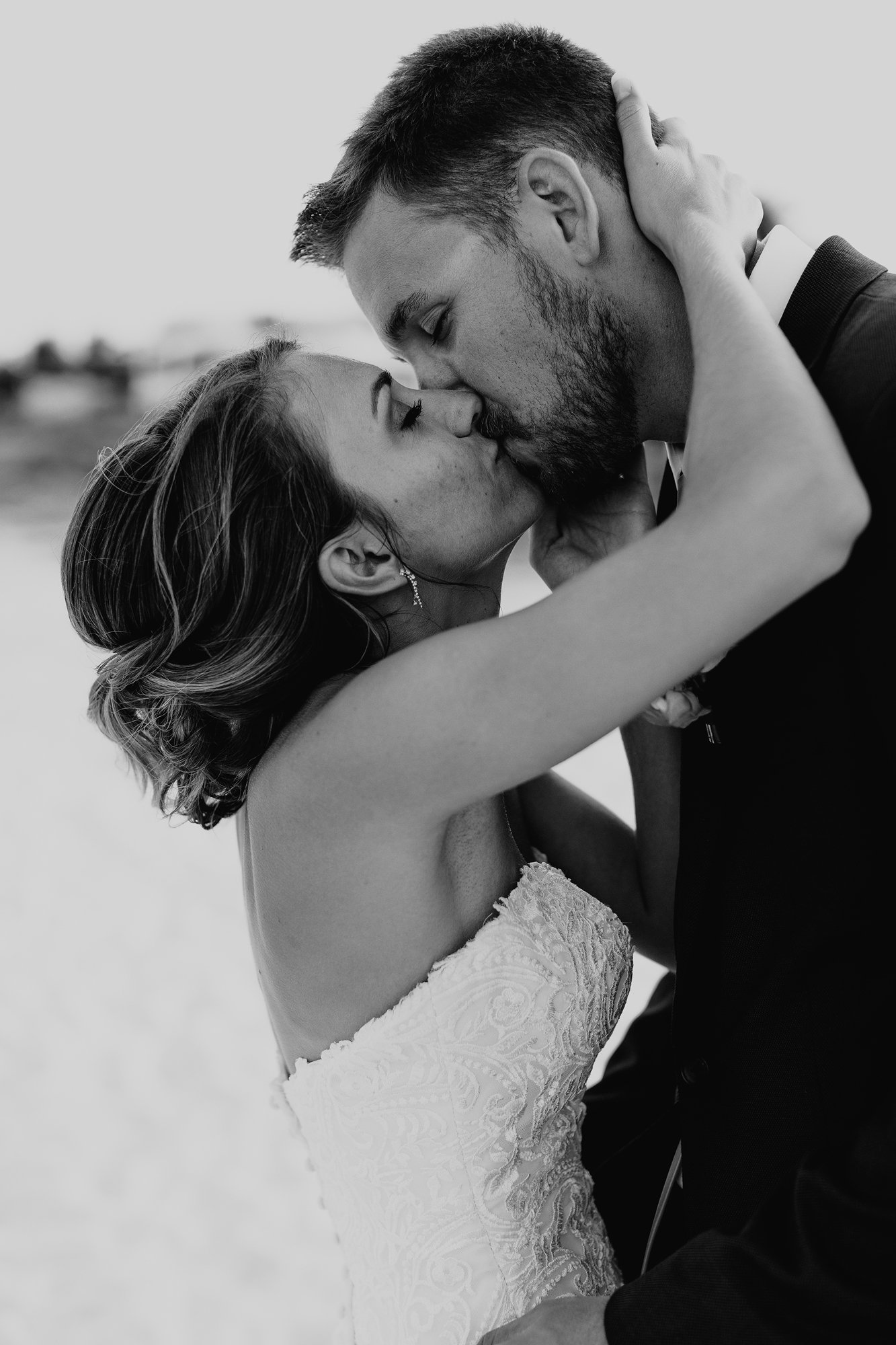 A black and white photo of a bride and groom kissing during their wedding, with the bride in a lace dress and the groom in a suit, outdoors on a cloudy day.