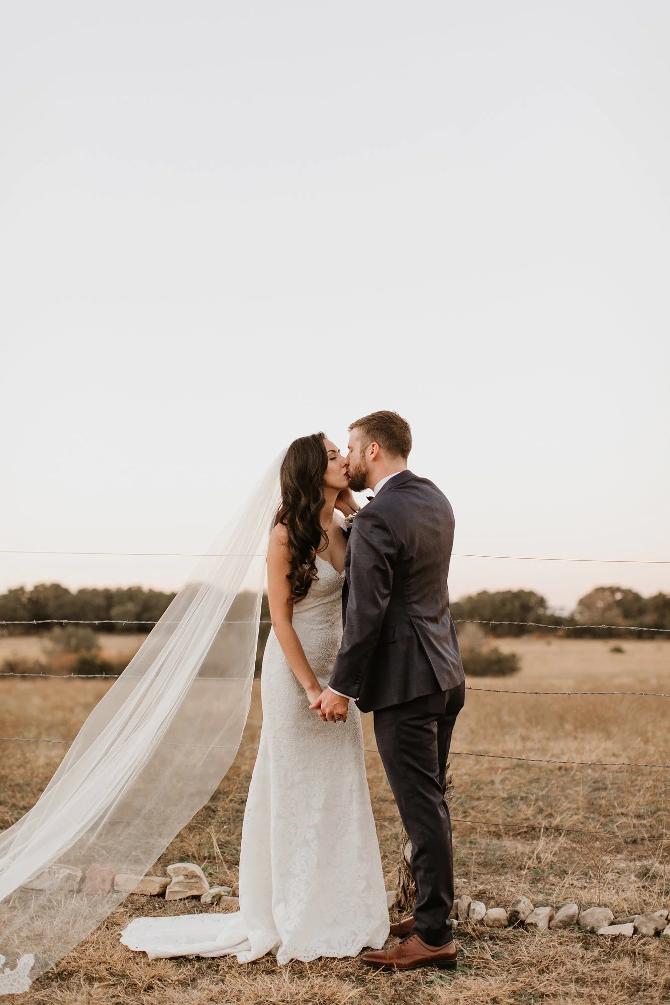 A bride and groom sharing a kiss and holding hands outdoors, with a barbed wire fence and open field in the background, during sunset or late afternoon.