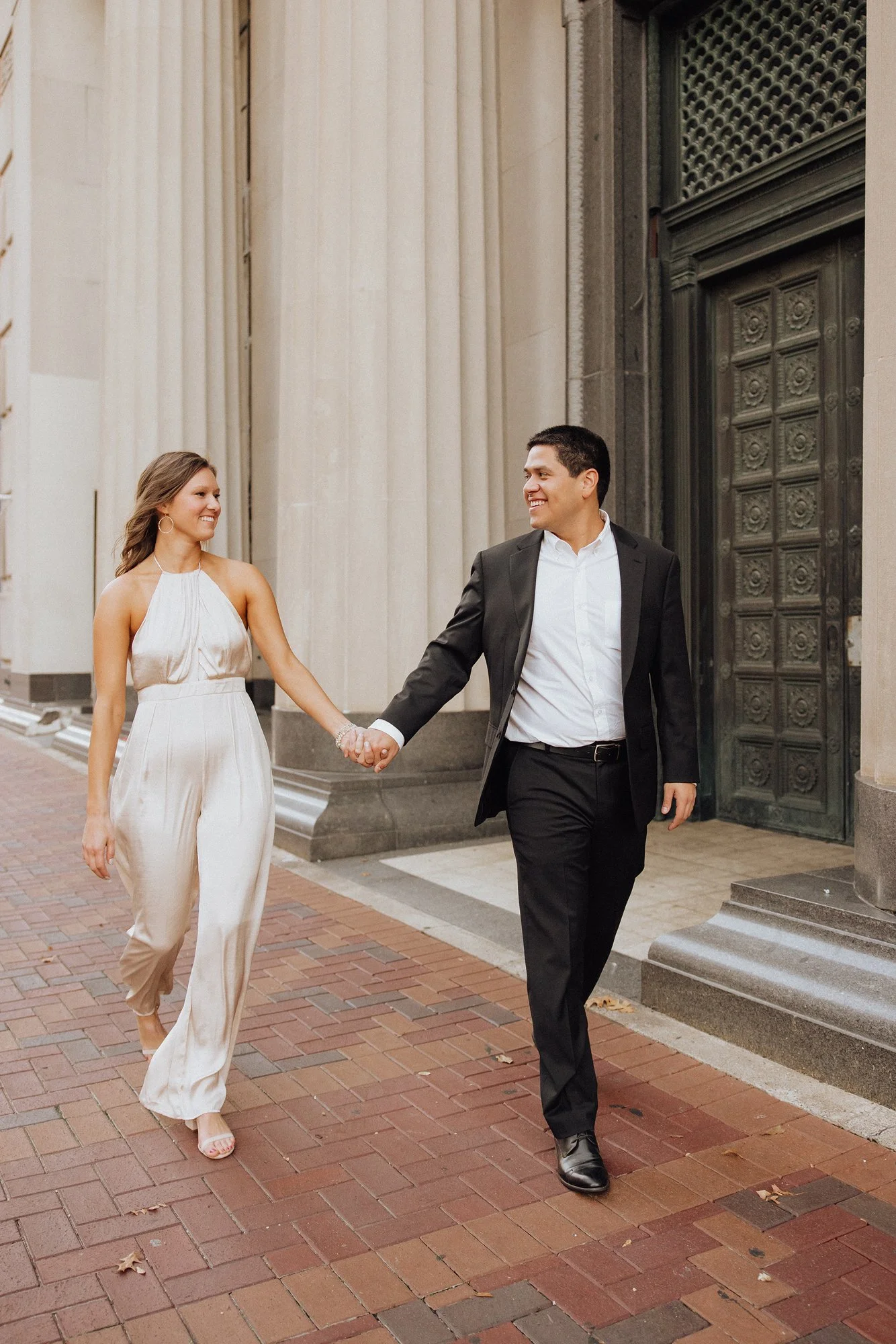 A couple, dressed in formal attire, walking hand in hand outside a historic building with large columns and ornate door, smiling at each other.