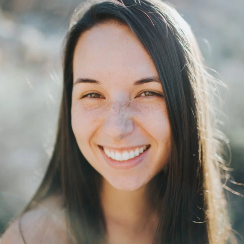 A young woman with long dark hair and freckles smiling outdoors on a sunny day.