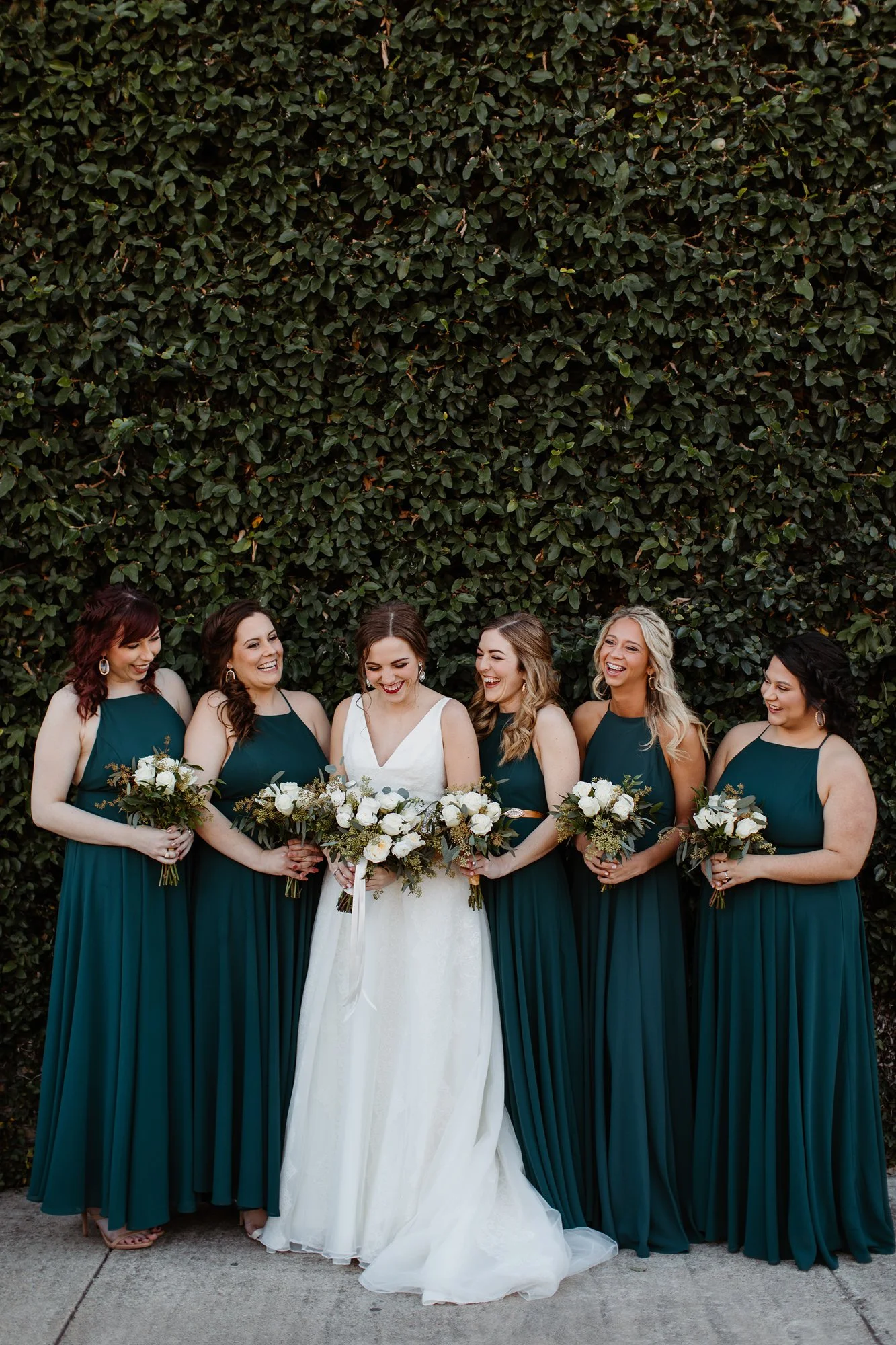 Group of seven women in formal dresses standing in front of a large green leafy hedge, holding bouquets of white flowers and greenery, smiling and laughing.