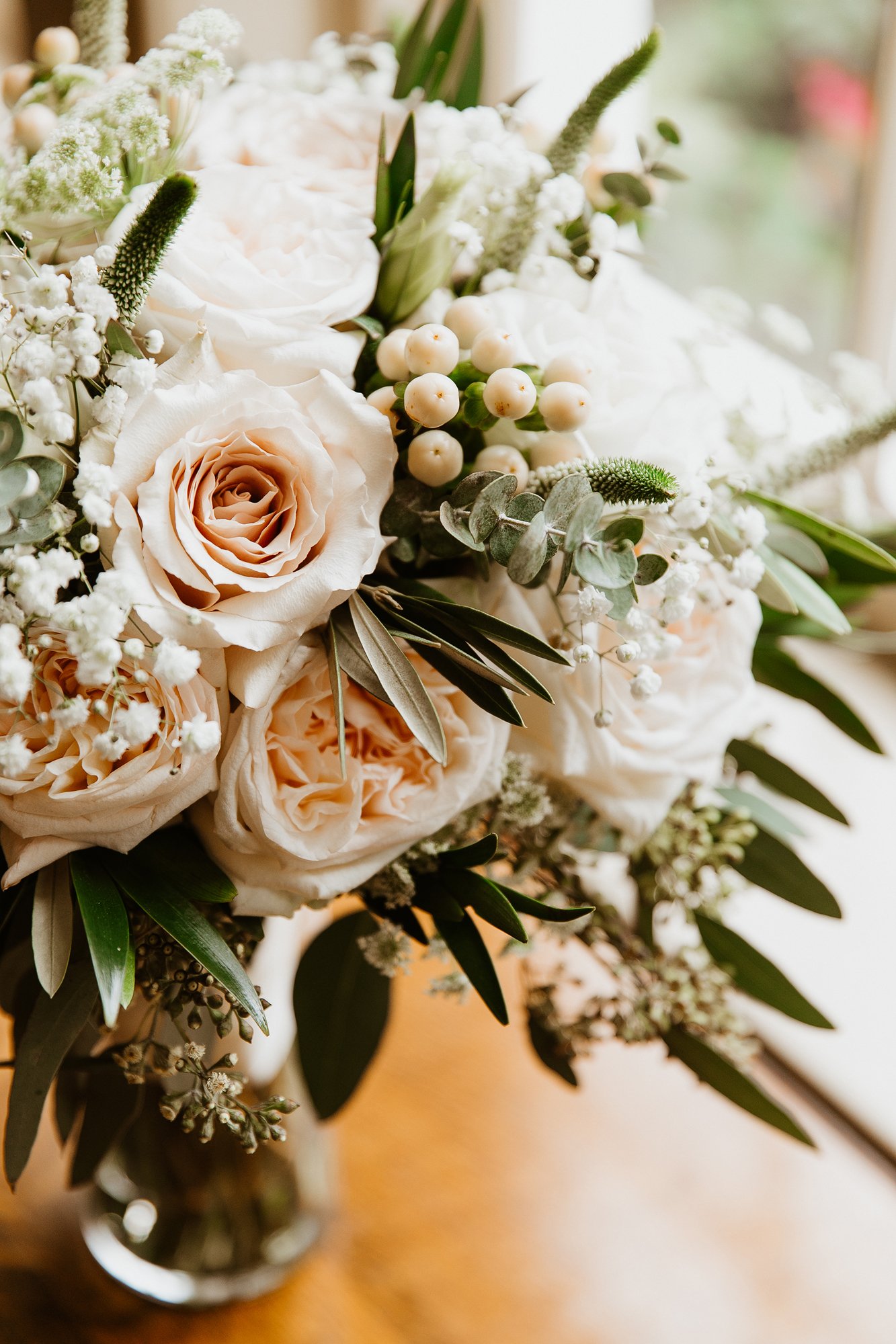 Close-up of a bouquet of white and pale peach roses with green foliage in a glass vase on a wooden surface.