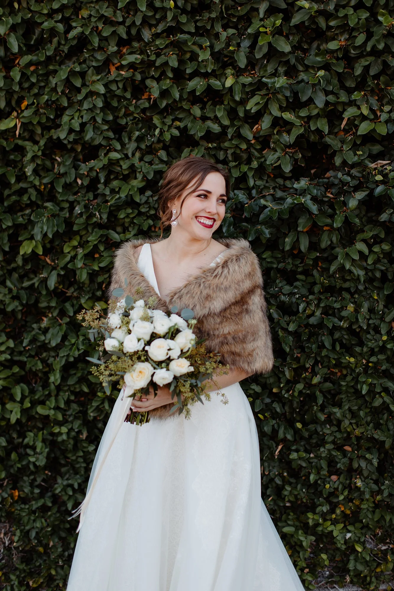 Joyful bride in a white wedding dress and fur wrap holding a bouquet of white flowers, standing in front of a green leafy hedge.