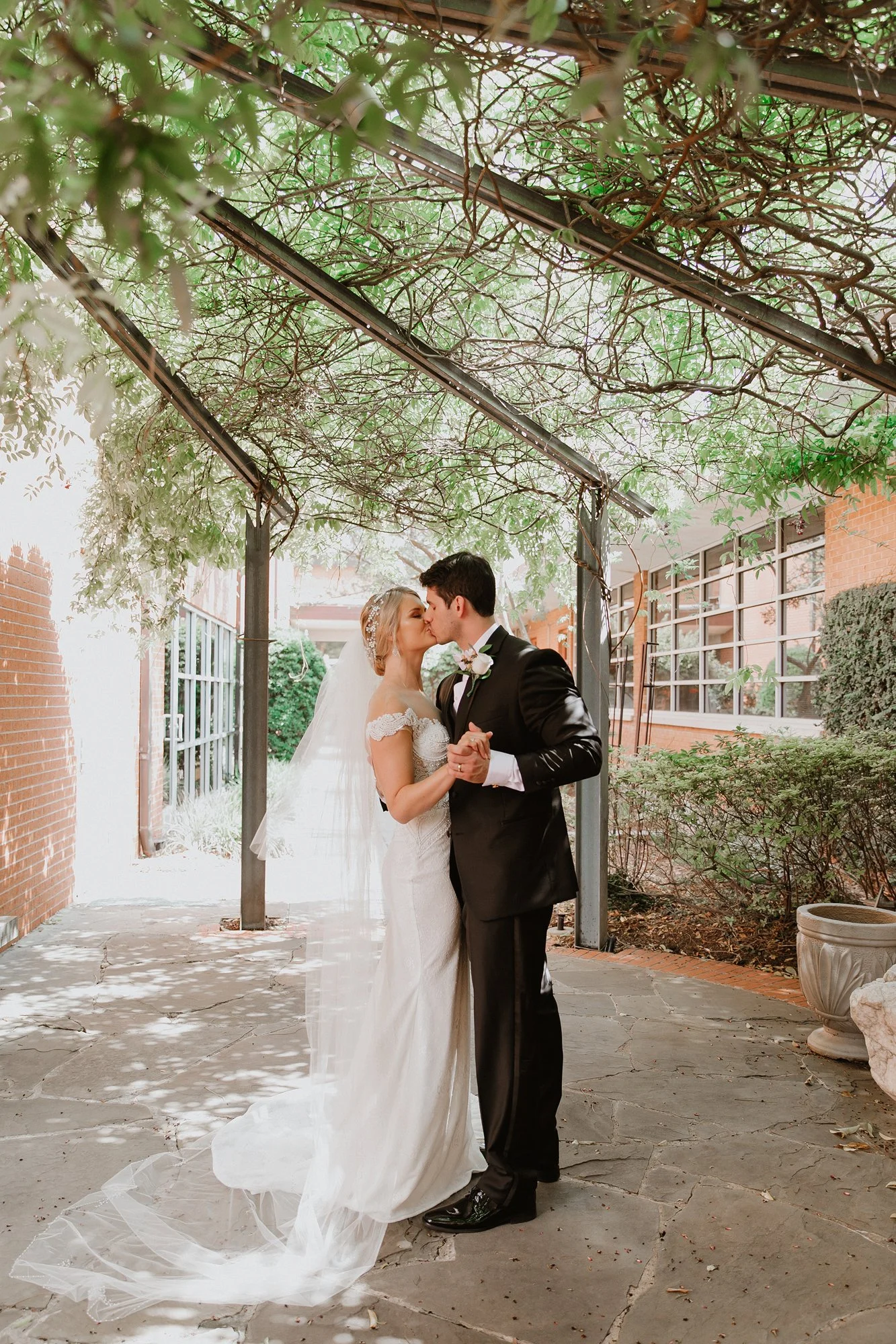 A bride and groom sharing a kiss and dancing under a leafy trellis in a garden setting, with brick buildings and large windows in the background.