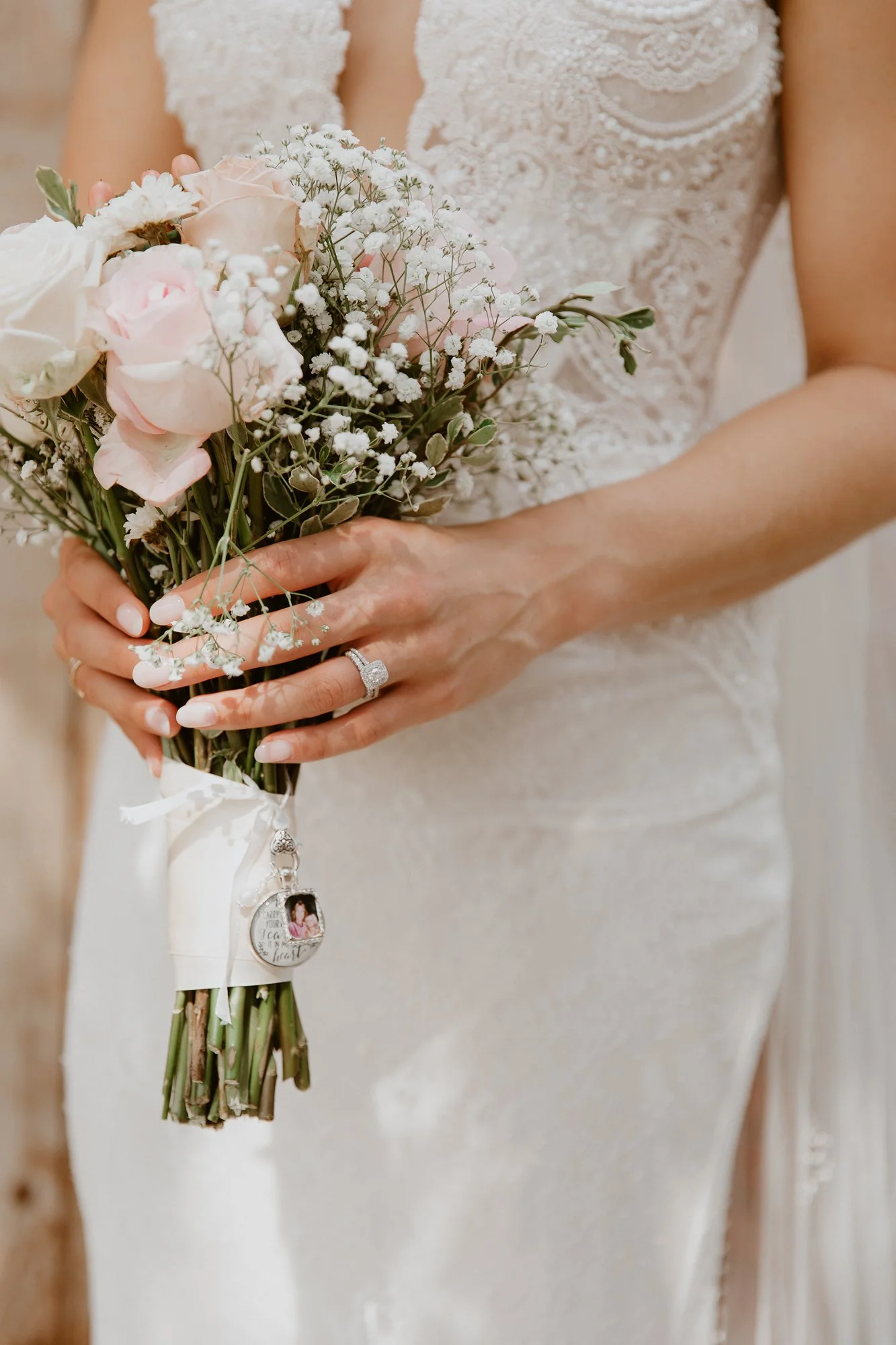 A woman in a wedding dress holding a bouquet of pink and white flowers. She is wearing rings and a wedding badge attached to her dress, with a bokeh background.