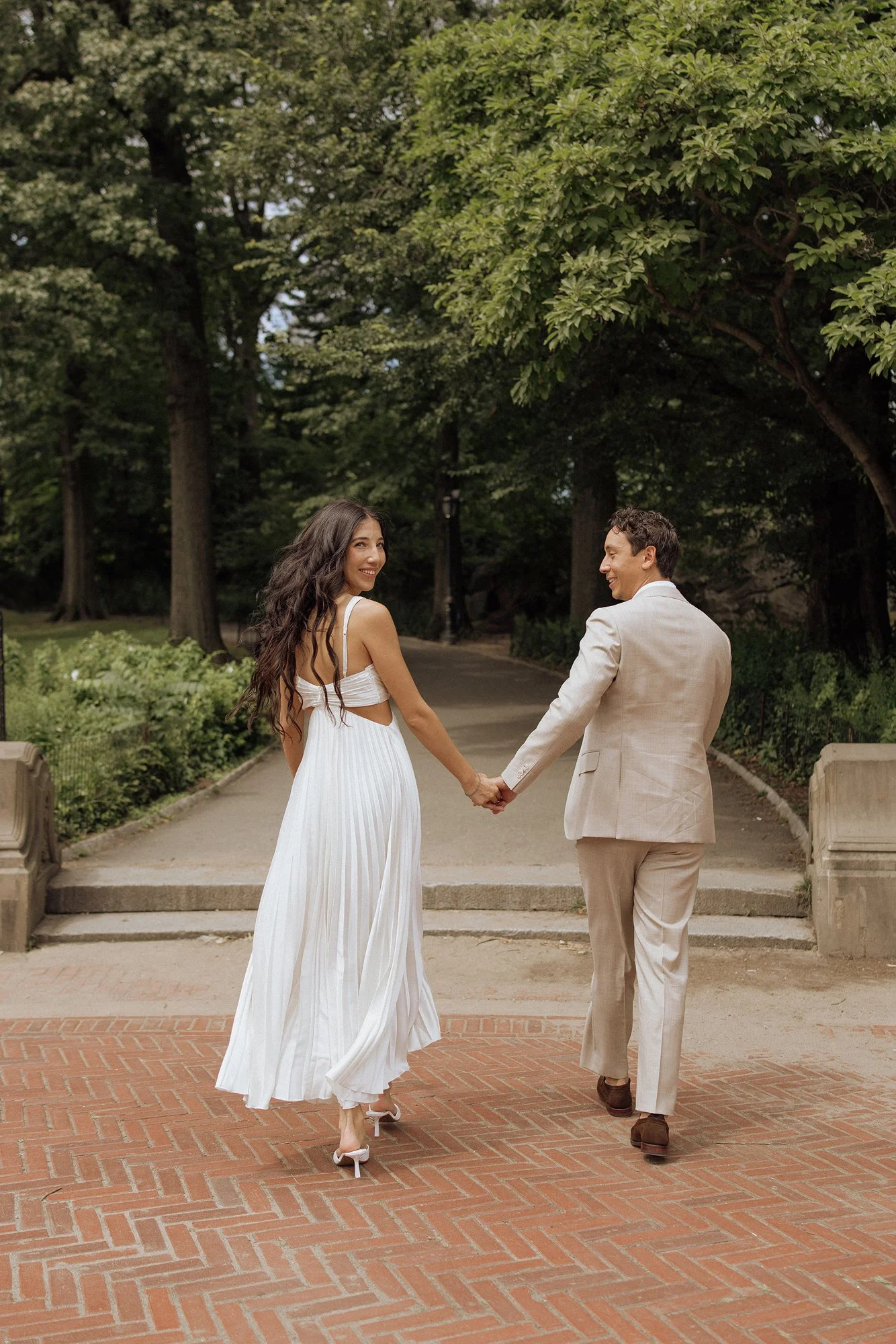A couple holding hands and walking in a park on a brick path, surrounded by trees. The woman is wearing a white dress and high heels, while the man is dressed in a light-colored suit.