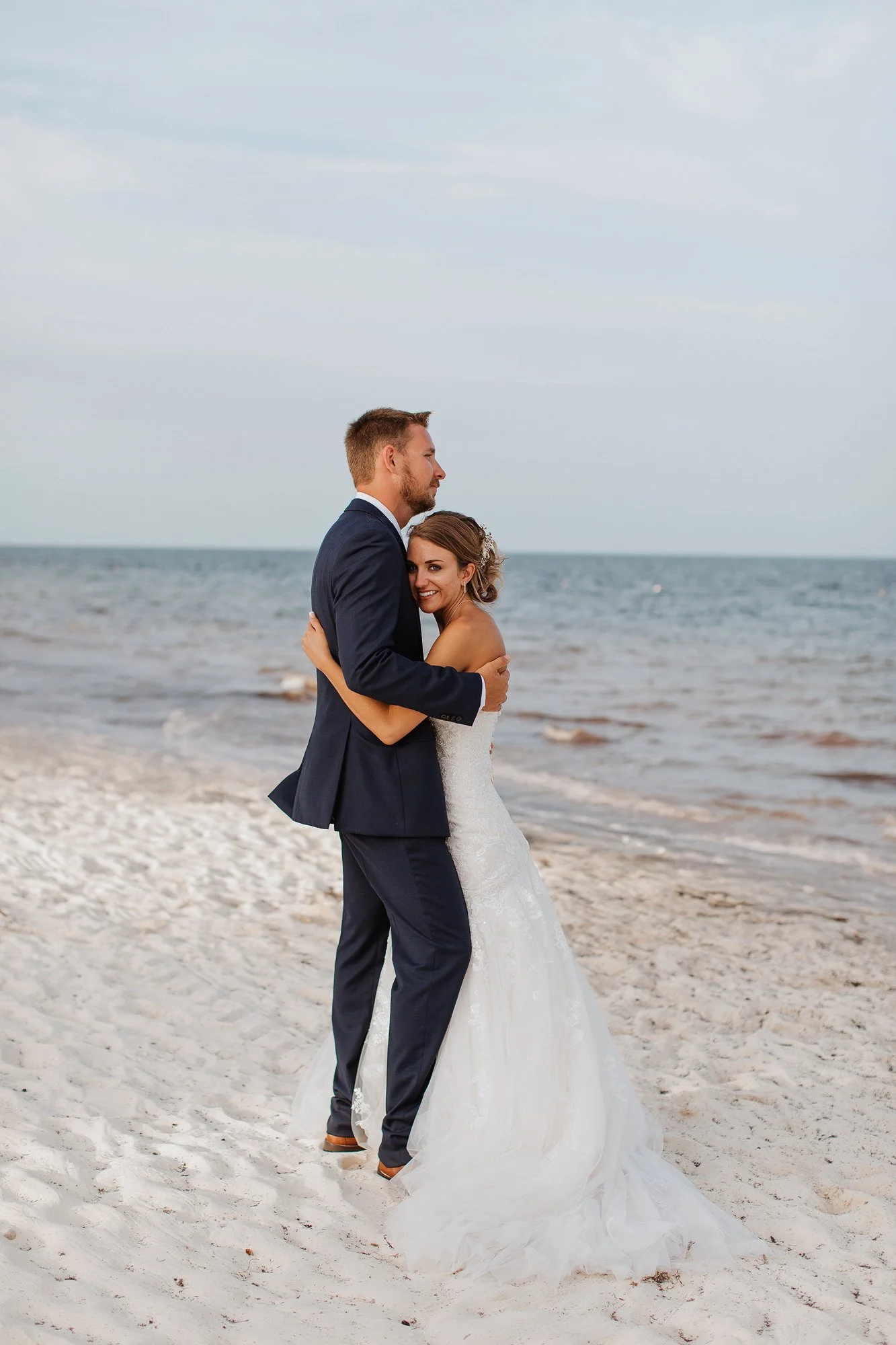 A newlywed couple embracing on a sandy beach with the ocean in the background. The man is in a dark suit and the woman is in a white wedding gown.