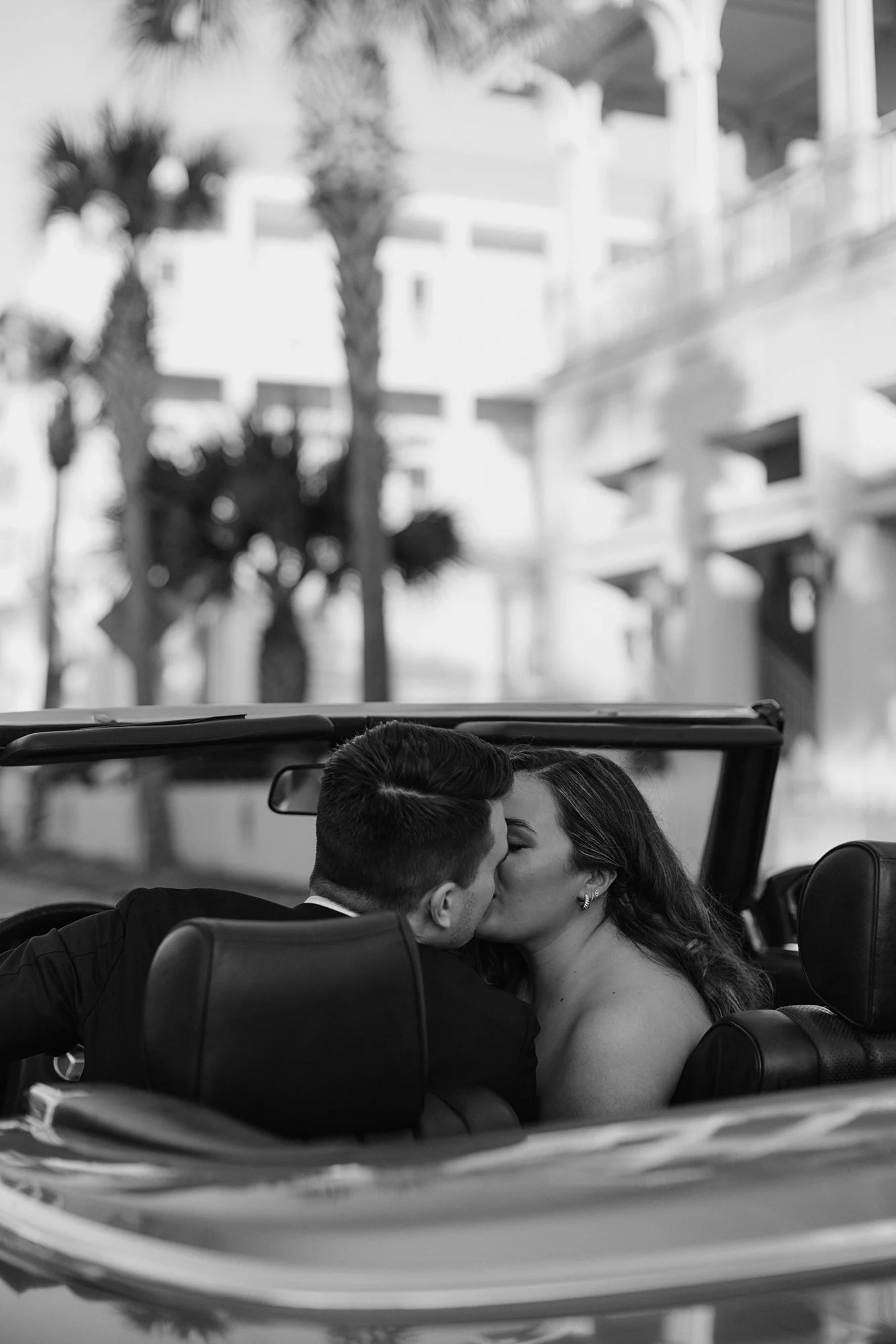 A couple sharing a kiss in a convertible car during daytime, with palm trees and apartment buildings visible in the background.