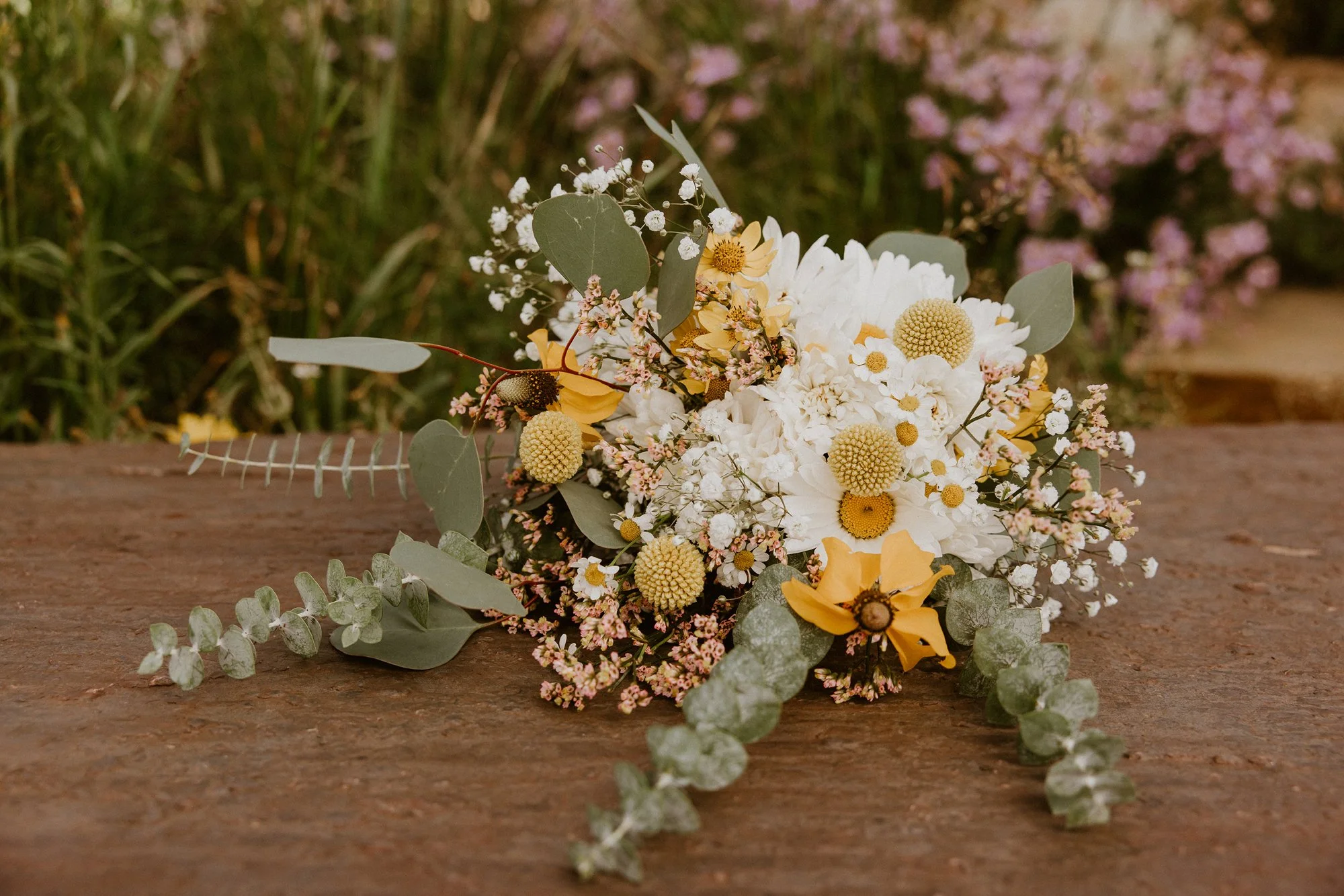 A flower arrangement with white, yellow, and pink flowers, eucalyptus leaves, and other greenery, placed on a wooden surface with a blurred green garden background.
