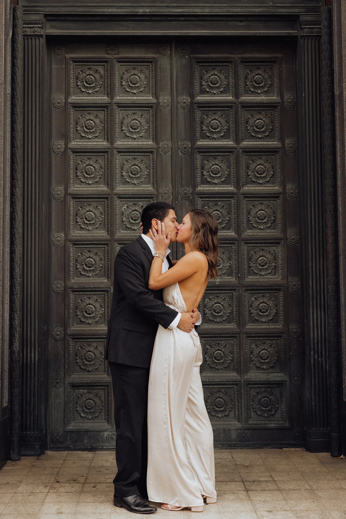A couple in wedding attire sharing a kiss in front of a large, ornate metal door.