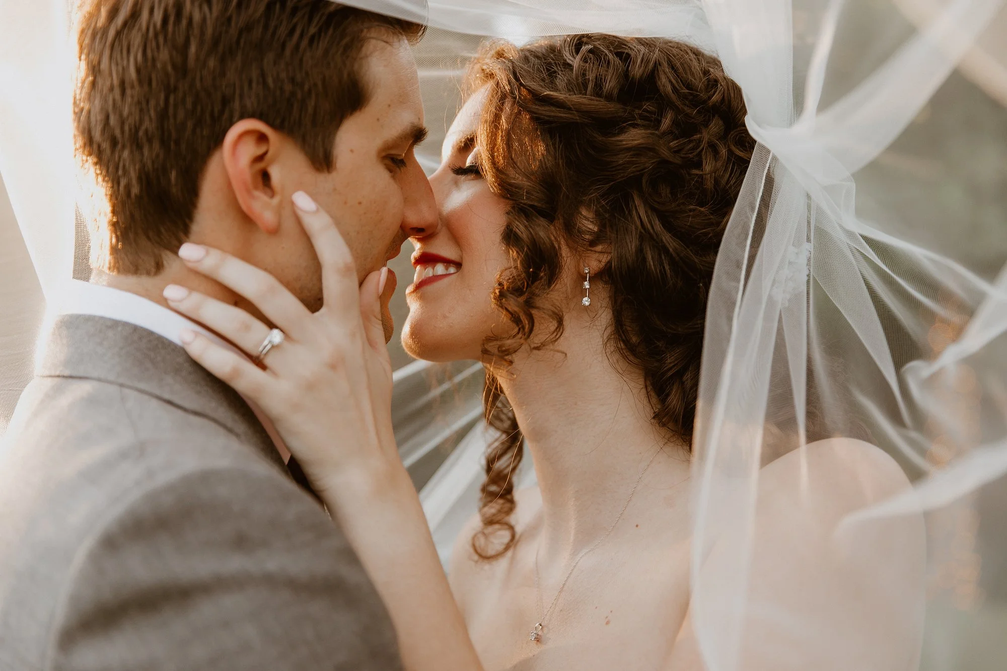 A couple sharing a romantic moment, close to each other, smiling with eyes closed, with gentle sunlight highlighting their faces. The woman wears earrings and a delicate necklace, with a sheer white veil draped around them.