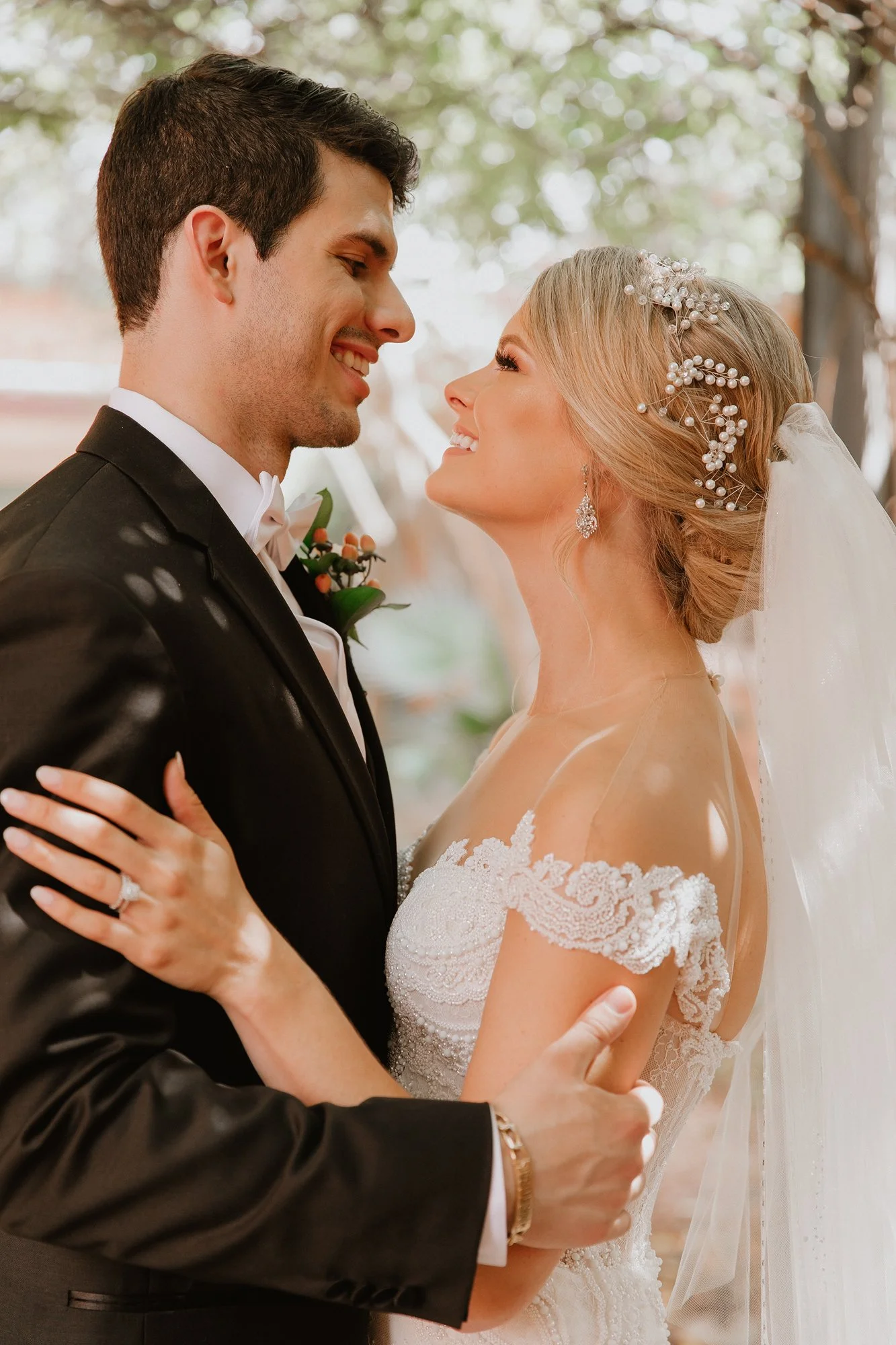A bride and groom smile at each other during their wedding, holding each other closely, with trees in the background.
