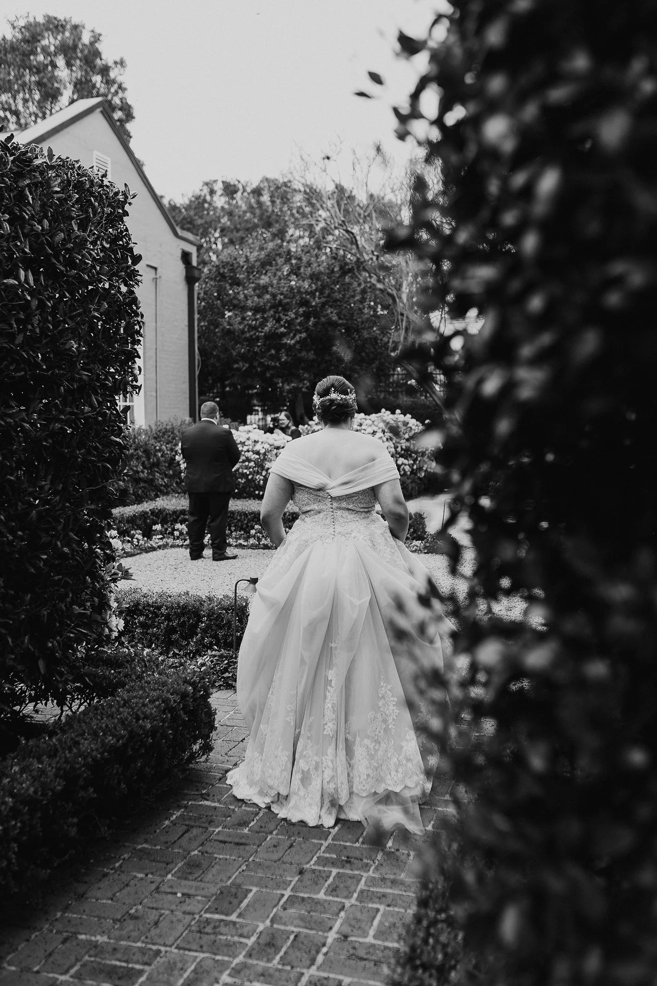 Black and white photo of a bride in a wedding gown walking along a brick pathway, with a man in a suit ahead of her, surrounded by bushes and trees, possibly outdoors during a wedding ceremony.