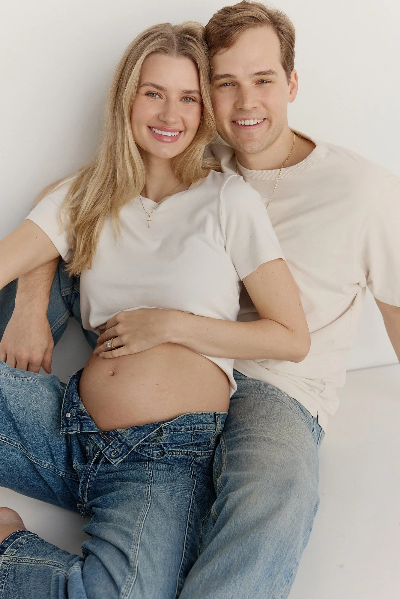 A smiling pregnant woman with long blond hair, wearing a white T-shirt and jeans, sitting on the lap of a man with short brown hair, dressed in a white T-shirt and jeans, both leaning against a plain white wall.