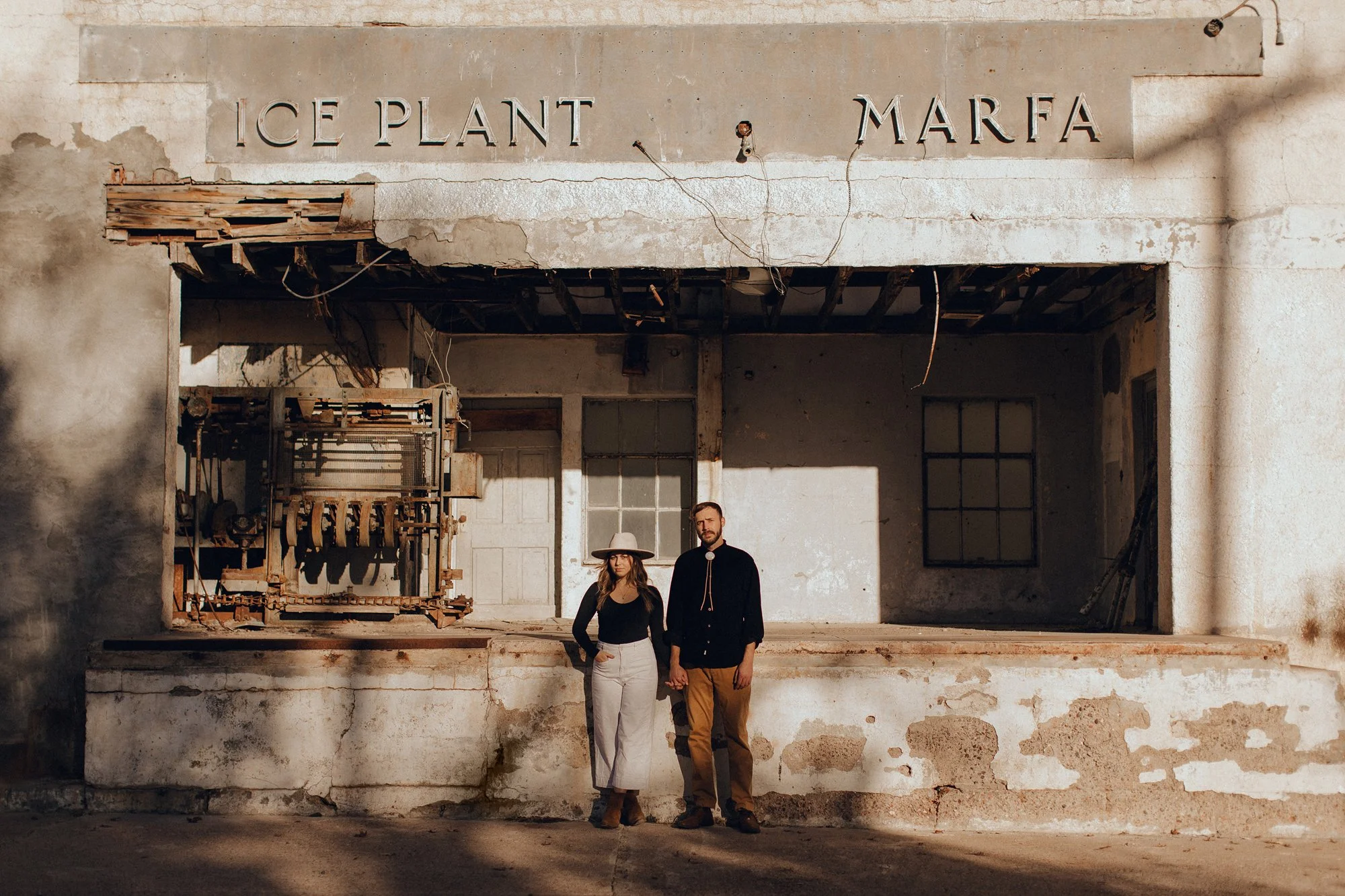 A man and a woman walking hand in hand in front of an abandoned building with a sign that reads 'Ice Plant Marfa.' The building has a weathered, peeling facade with exposed brick and old equipment inside. The sunlight casts shadows on the scene.