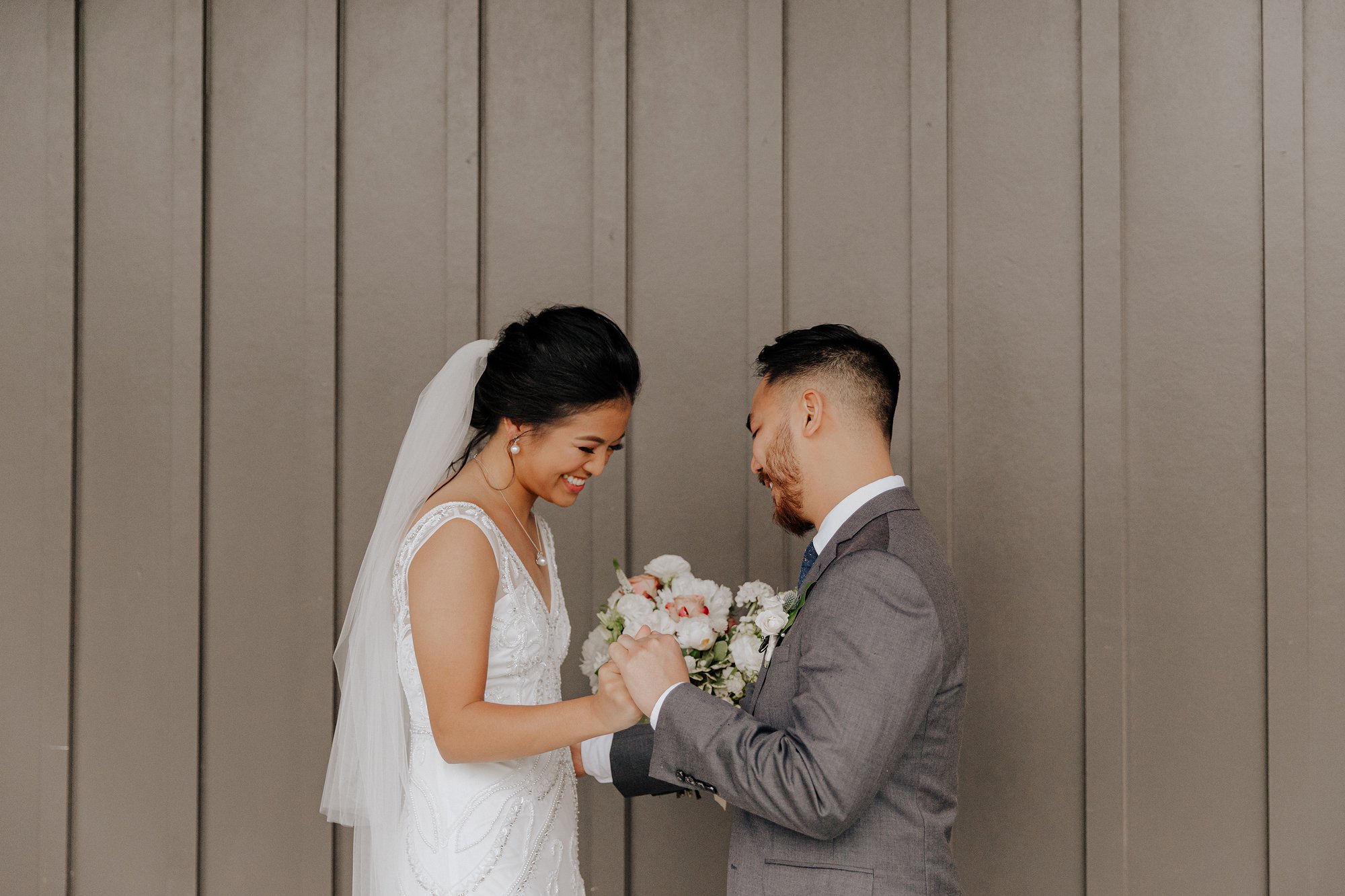 A bride and groom stand facing each other during their wedding ceremony, holding hands, with the bride smiling and holding a bouquet of flowers.