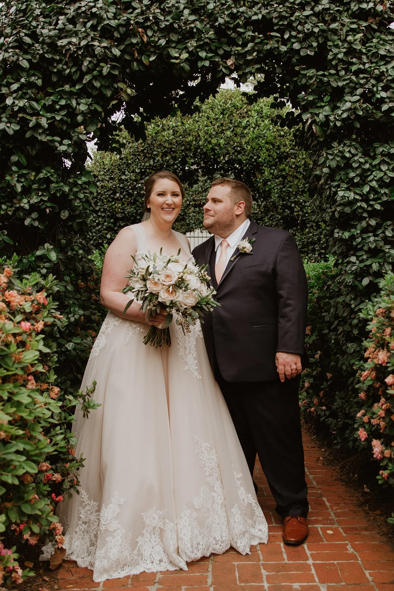 A bride in a white wedding gown holding a bouquet of roses and greenery, standing with a groom in a black suit and white shirt, under a leafy archway during a wedding.