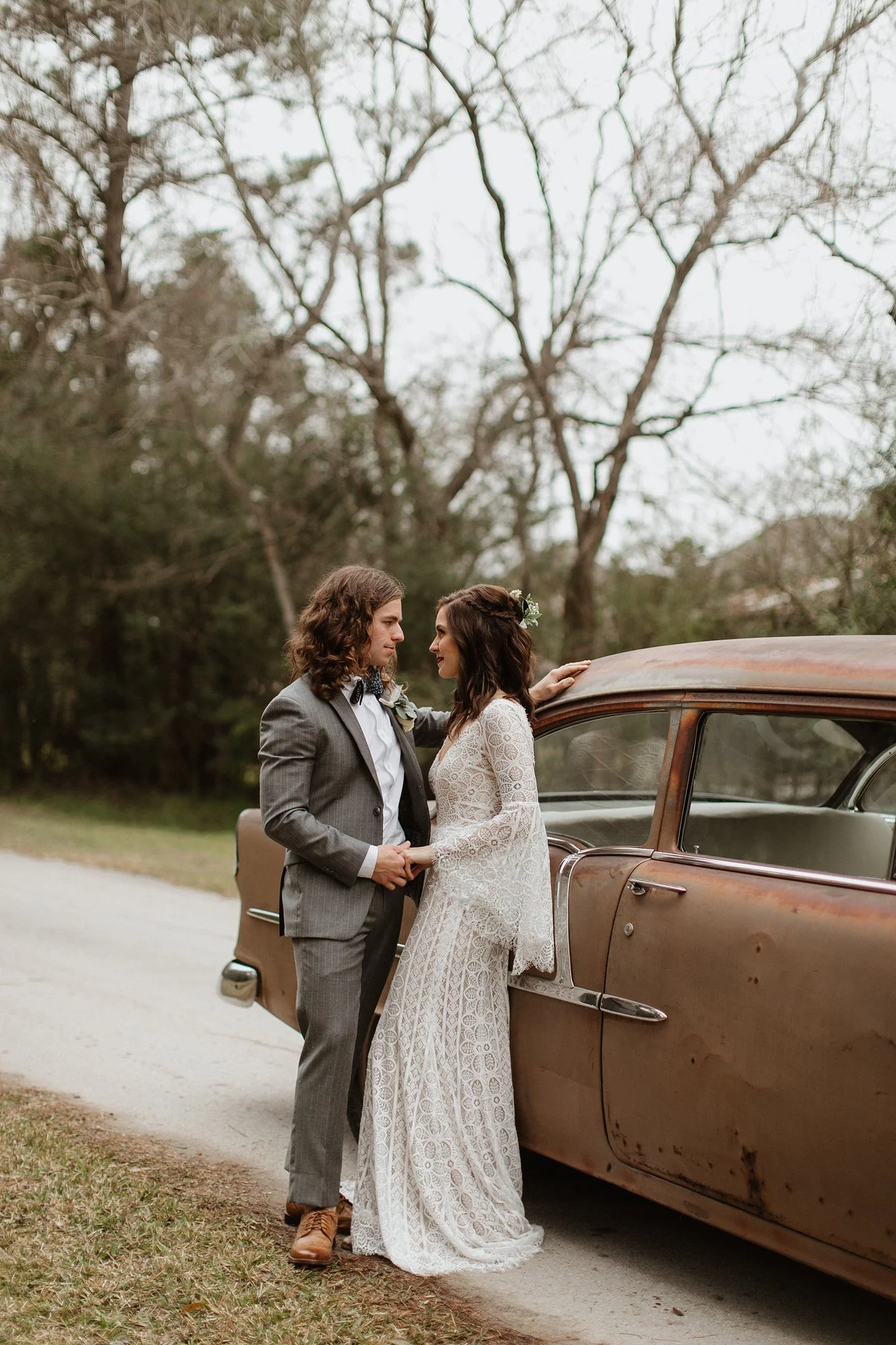 A couple dressed in vintage wedding attire standing close to each other next to an old-fashioned car outdoors, with leafless trees in the background.