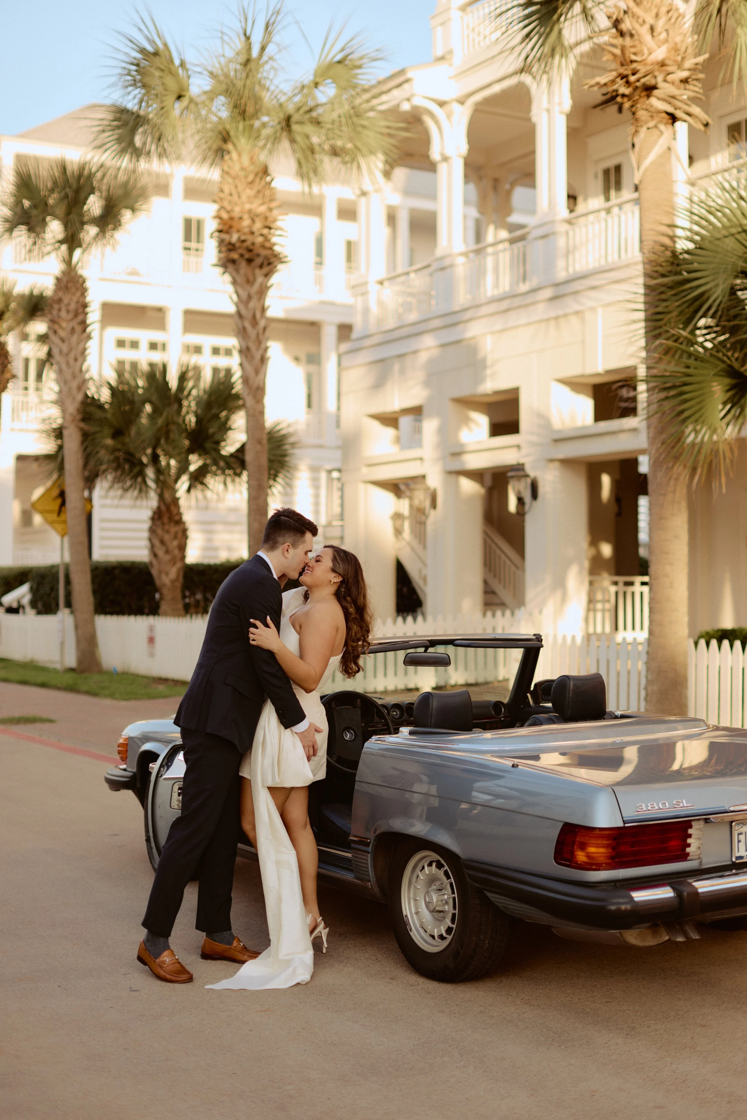 A couple dressed in wedding attire standing next to a gray convertible car parked on a street with palm trees and white residential buildings in the background. The man is in a suit, and the woman is in a white dress, both smiling and close to each o