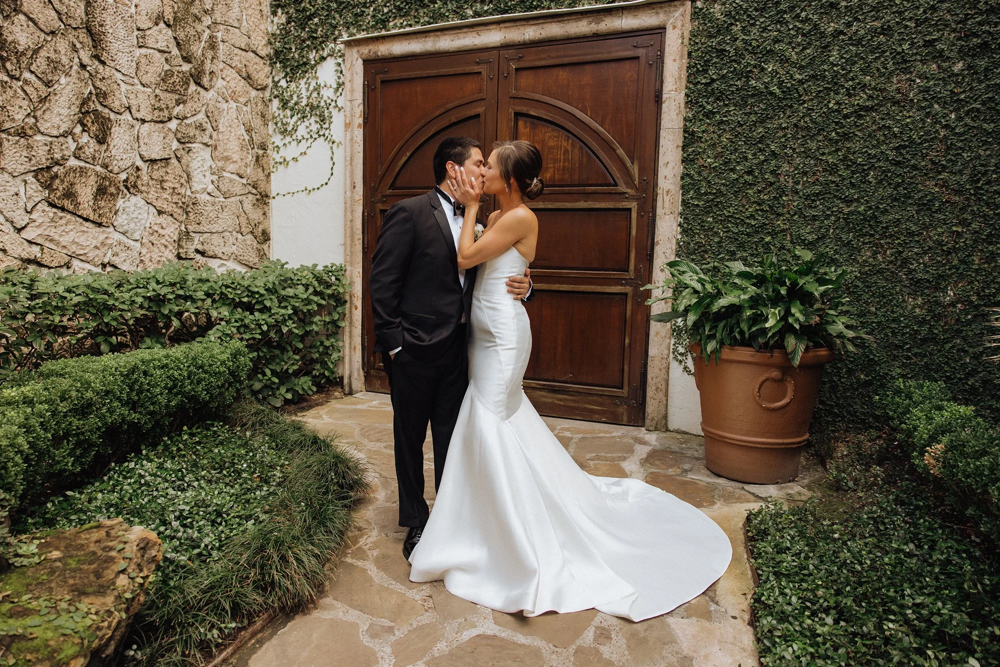A bride and groom sharing a kiss in front of a wooden door surrounded by greenery and stone walls.