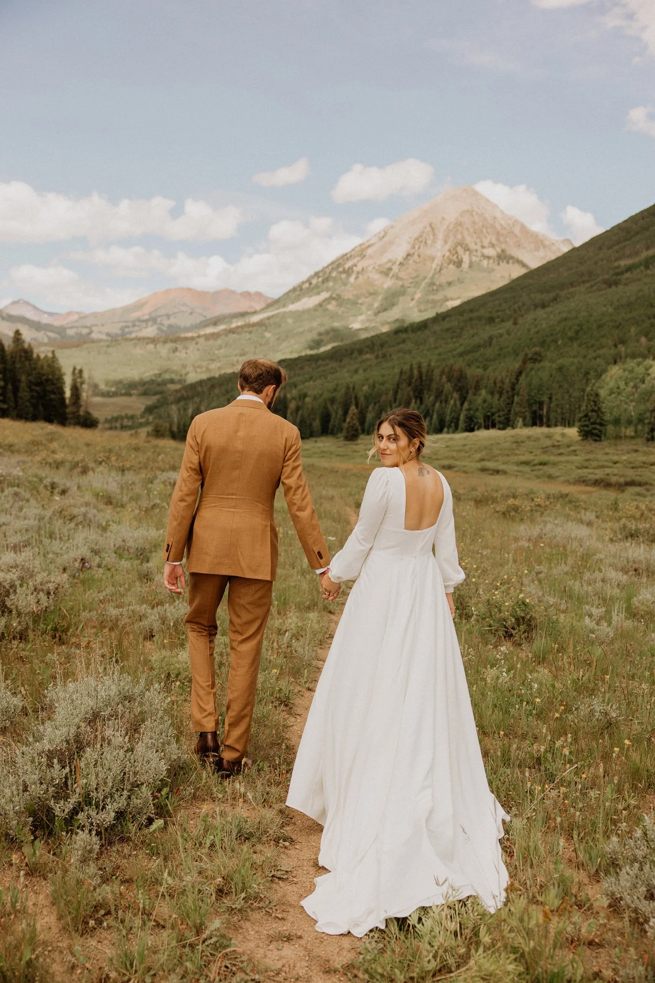 A man and woman holding hands walking through a grassy field in a mountainous landscape, with the woman wearing a white wedding dress and the man in a tan suit, under a sky with scattered clouds.
