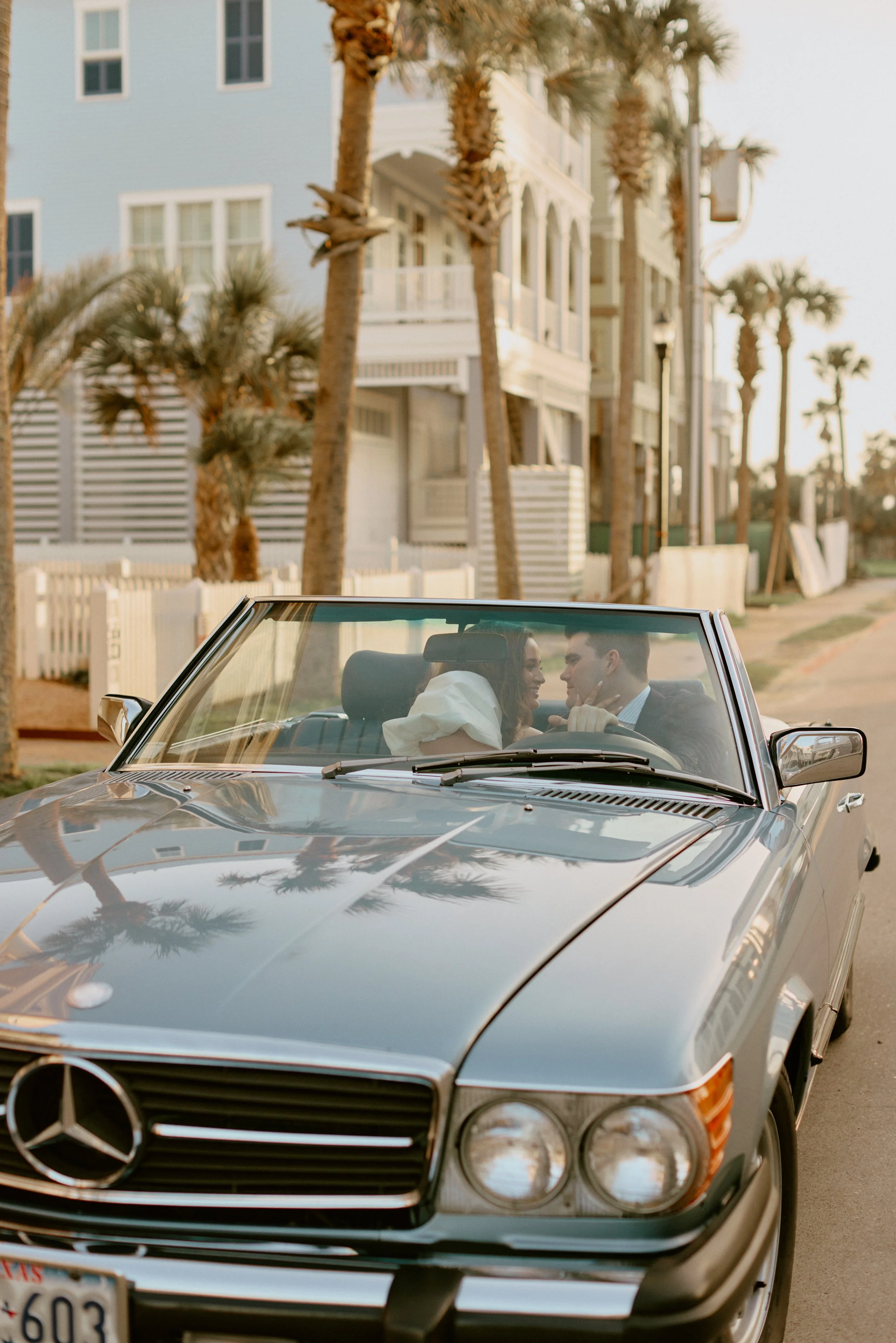 A smiling woman and man are sitting in a vintage black Mercedes-Benz convertible, facing each other, with palm trees and beach houses in the background.