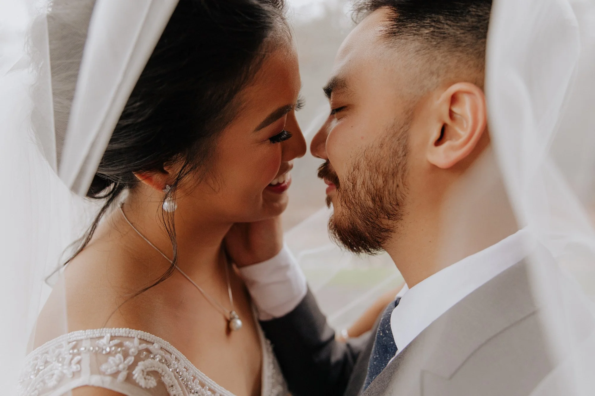 A couple is inside a white, semi-transparent fabric tent, with their faces close and smiling at each other. The woman has dark hair, earrings, and a white dress with embroidery, while the man has short dark hair, a beard, and is wearing a suit with a