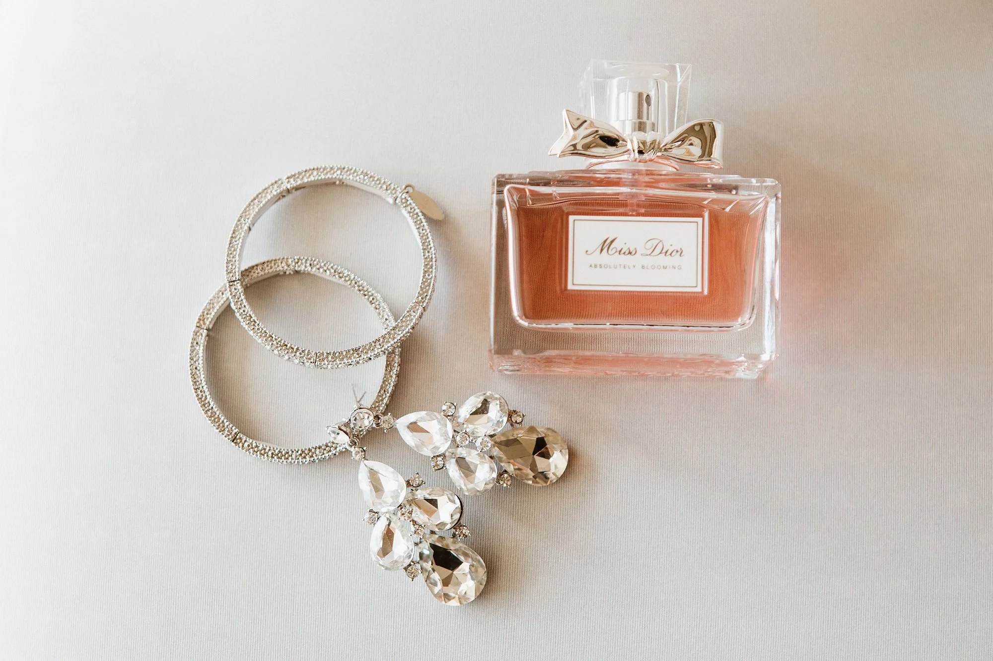 A collection of jewelry including diamond and pearl earrings, a pearl bracelet, and an ornate necklace, alongside a bottle of Miss Dior perfume with a bow on the cap, on a white surface.